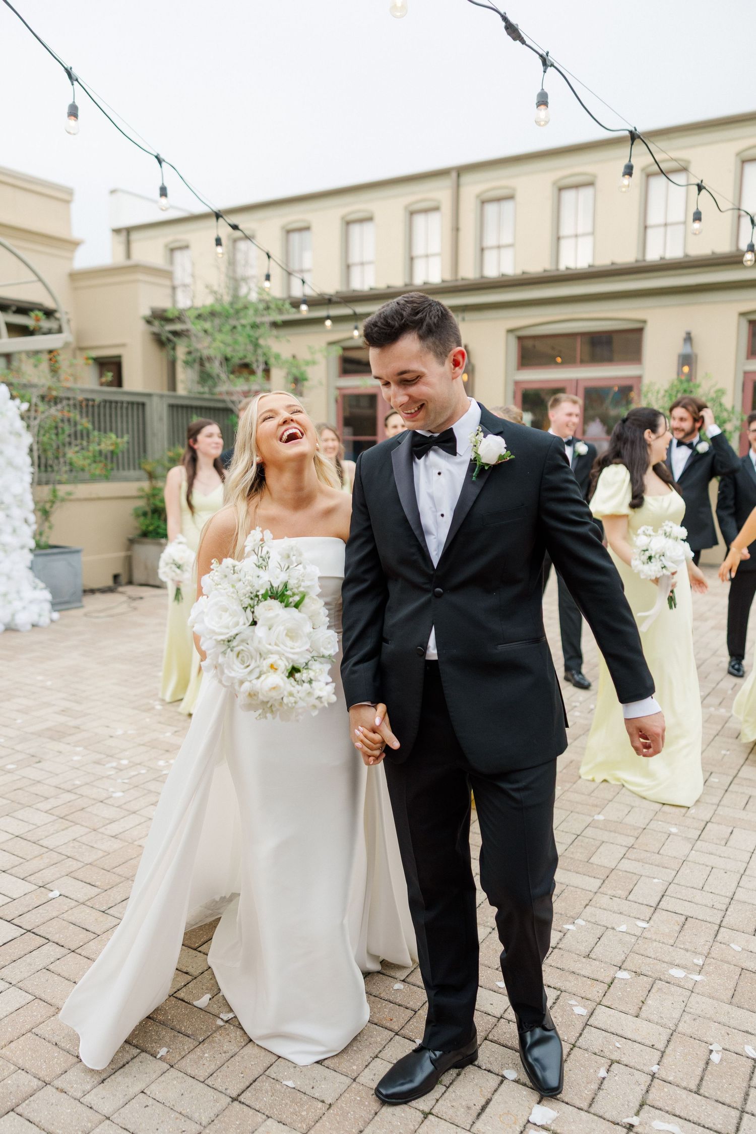Couple in formal wedding attire dancing together on an outdoor stone patio with guests in background.