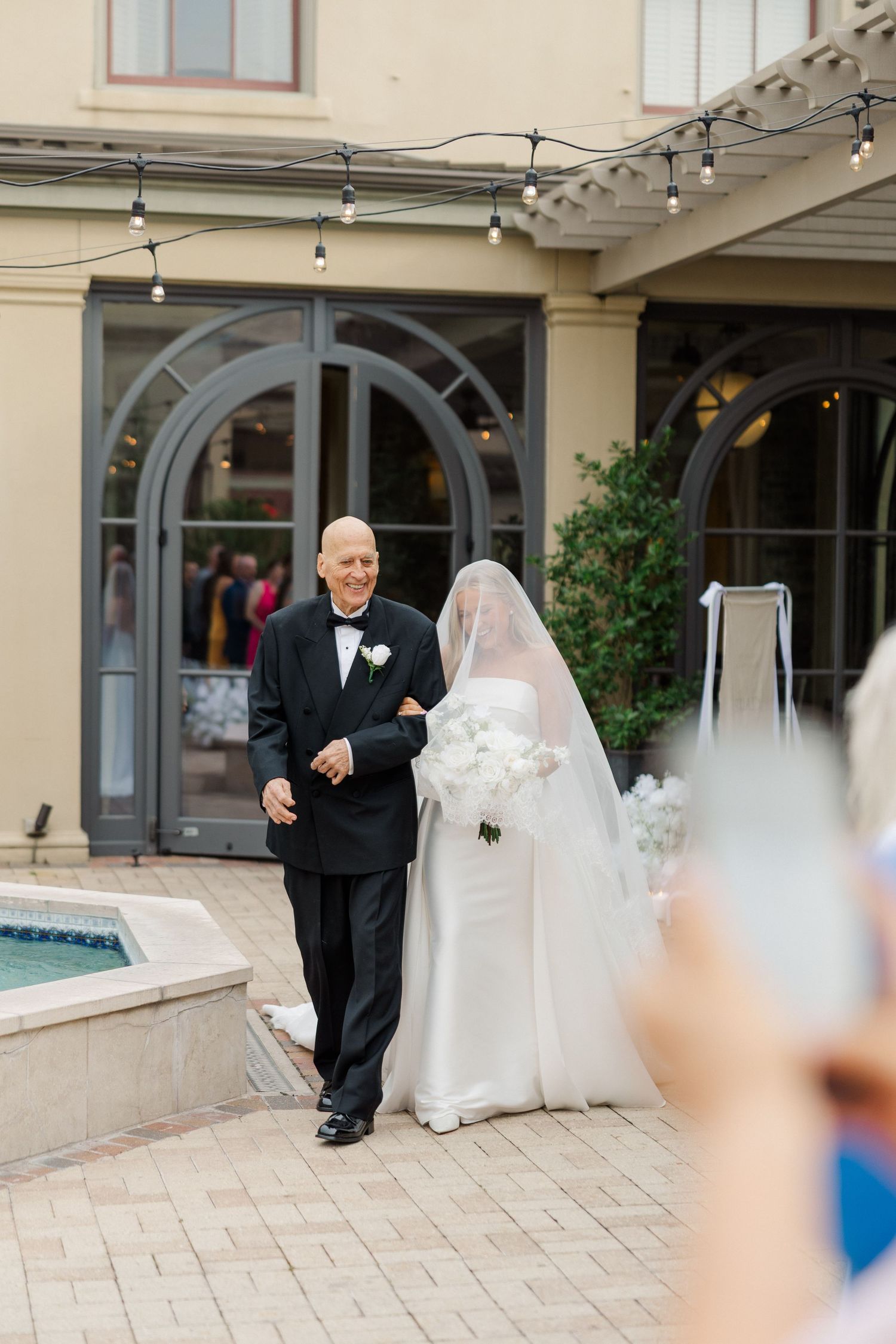 A wedding party walks along a paved outdoor courtyard with elegant arched doorways.