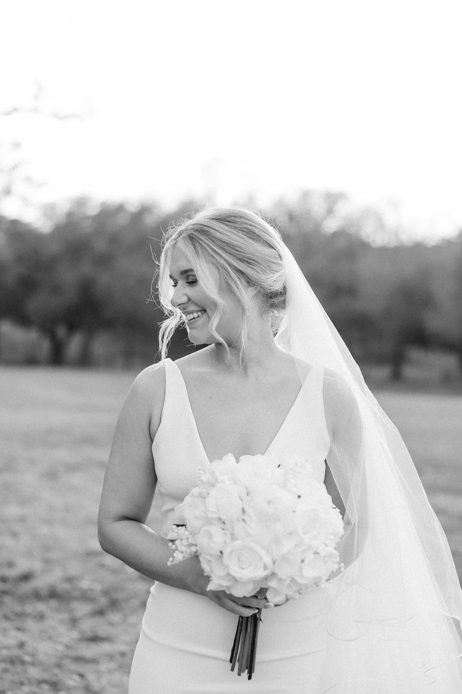 A joyful bride in a simple white v-neck wedding dress holds white bouquet while posing outdoors.