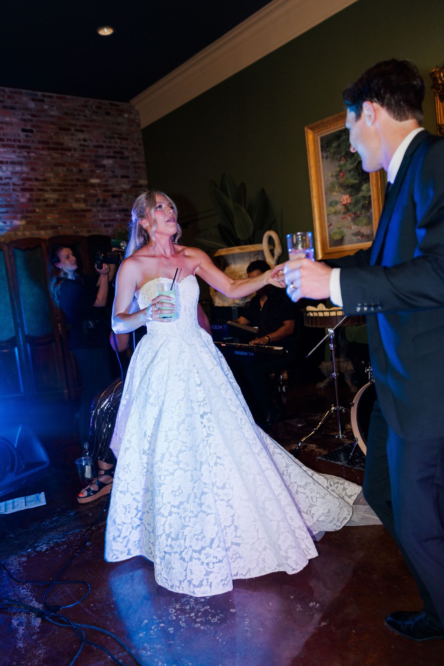 Bride in strapless lace ballgown dances with groom while toasting champagne at lively wedding reception.
