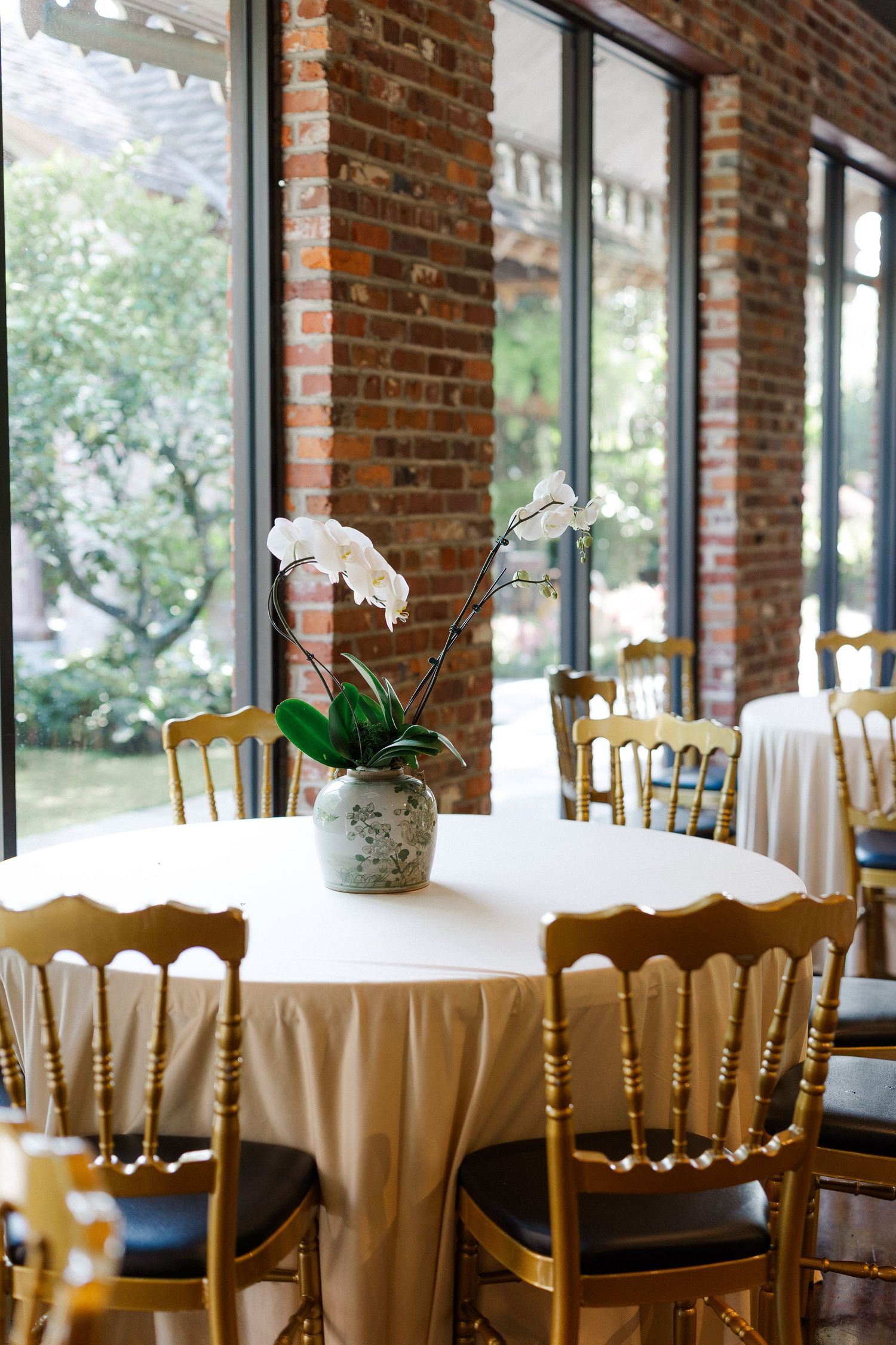 Elegant wedding reception table with white orchid centerpiece, gold chiavari chairs, and white linen in a brick-walled venue.