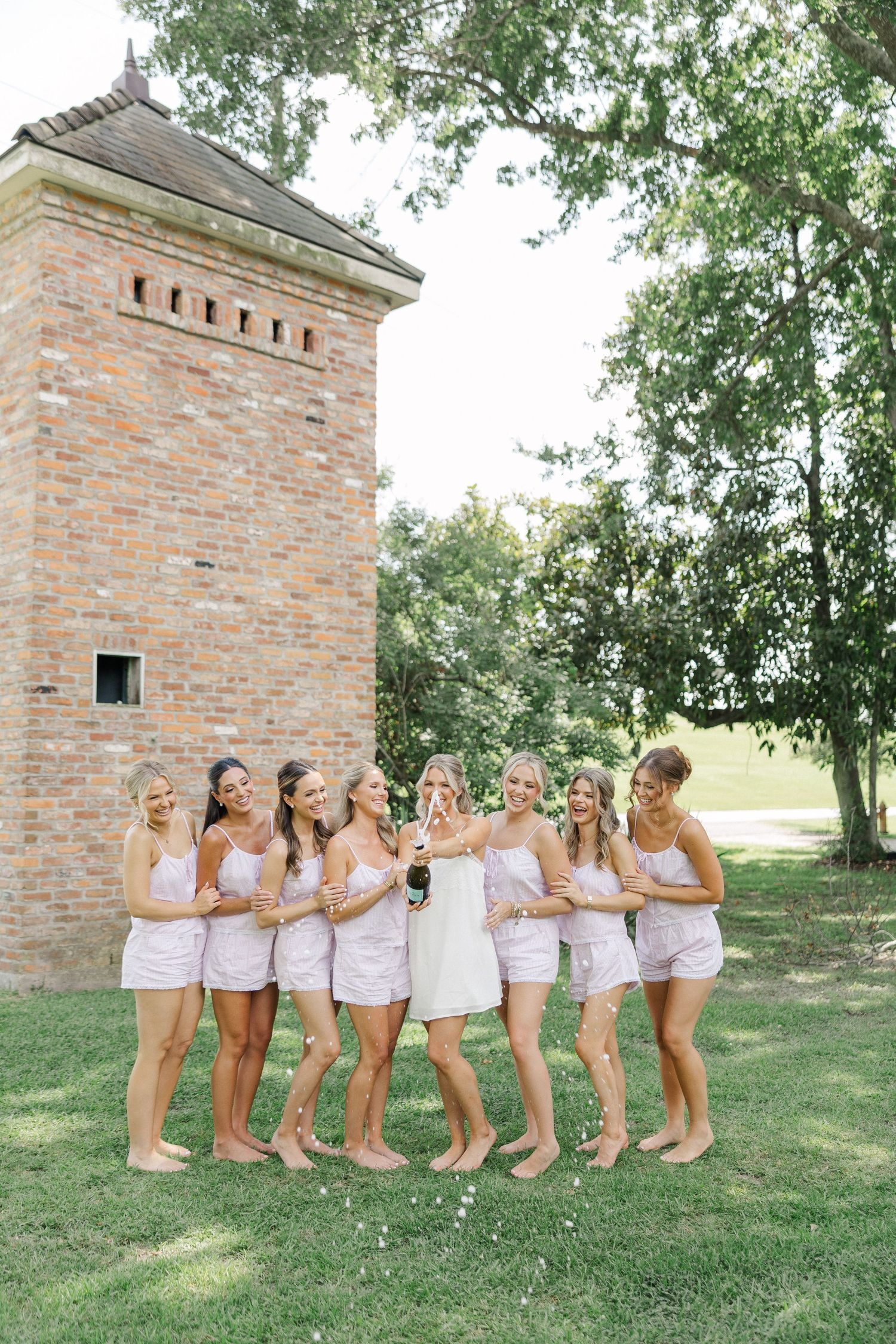 Bride and bridesmaids in matching pink pajamas celebrating with champagne near a brick tower outdoors.