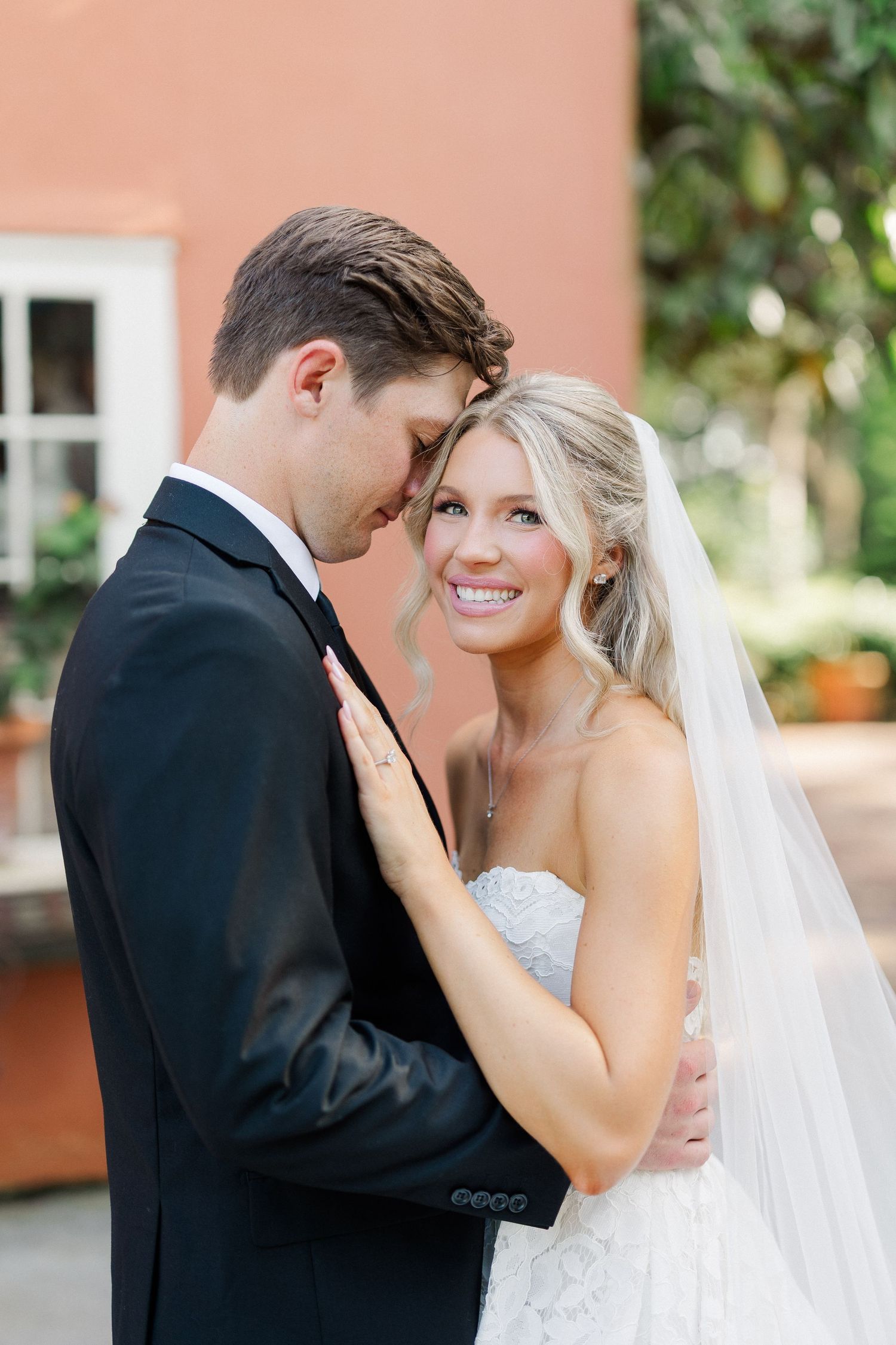 Bride in lace gown smiles joyfully at groom in black tuxedo during romantic wedding portrait.