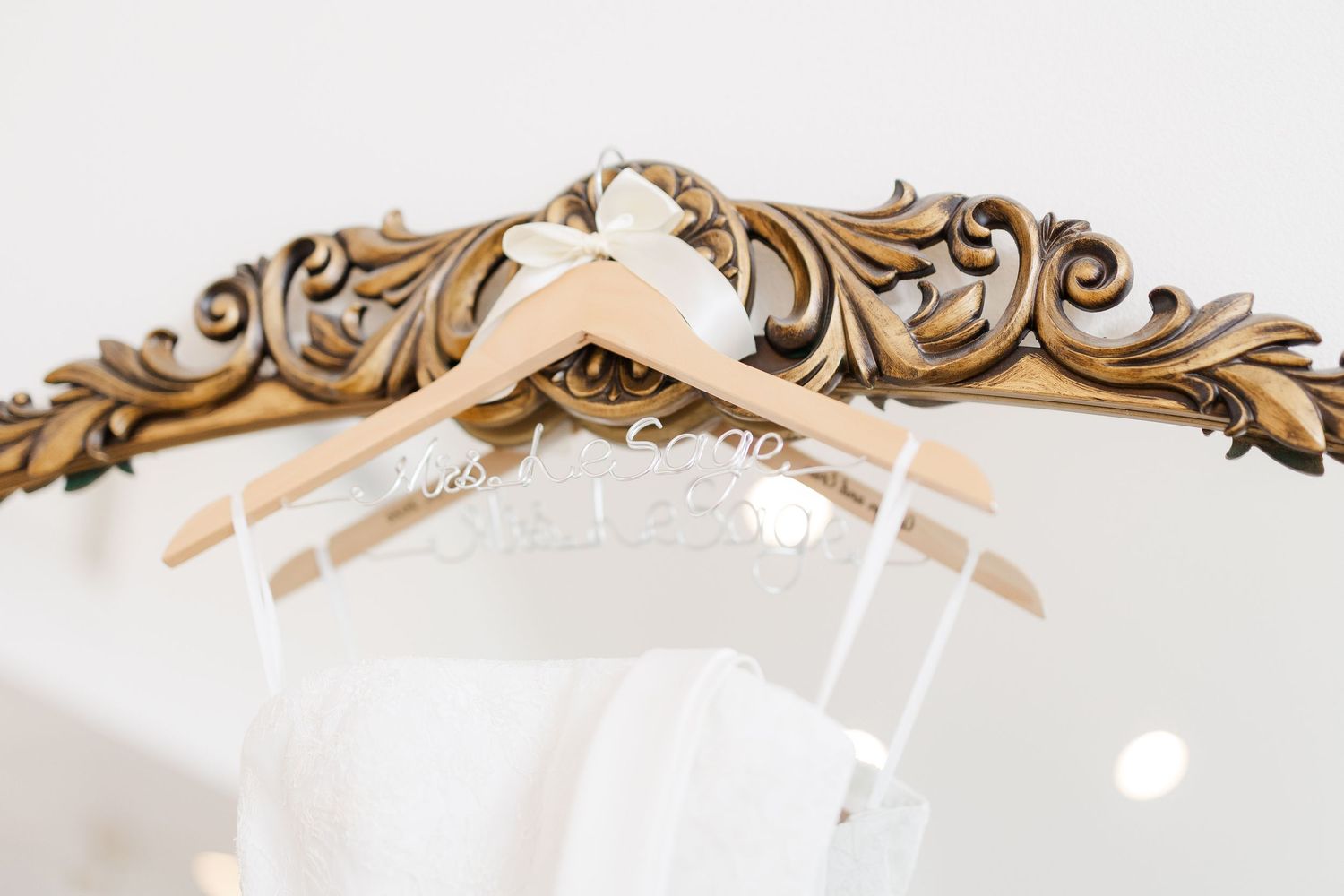 White wedding dress hanging on wooden hanger over ornate gold mirror frame.