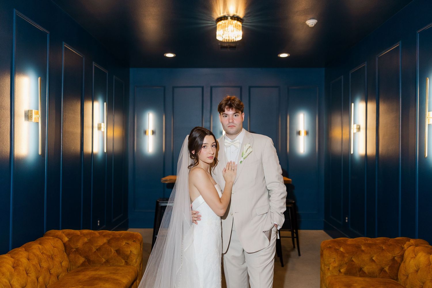 Bride and groom pose elegantly in a moody blue room with gold sconce lighting and velvet orange seating.