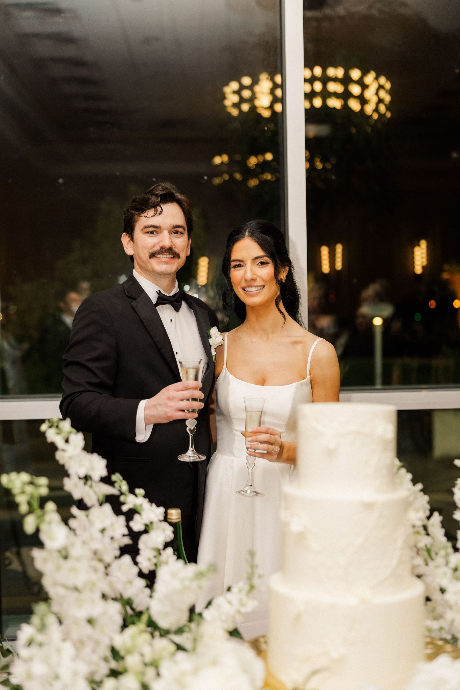 Bride and groom in wedding attire pose with champagne flutes beside elegant white tiered wedding cake.