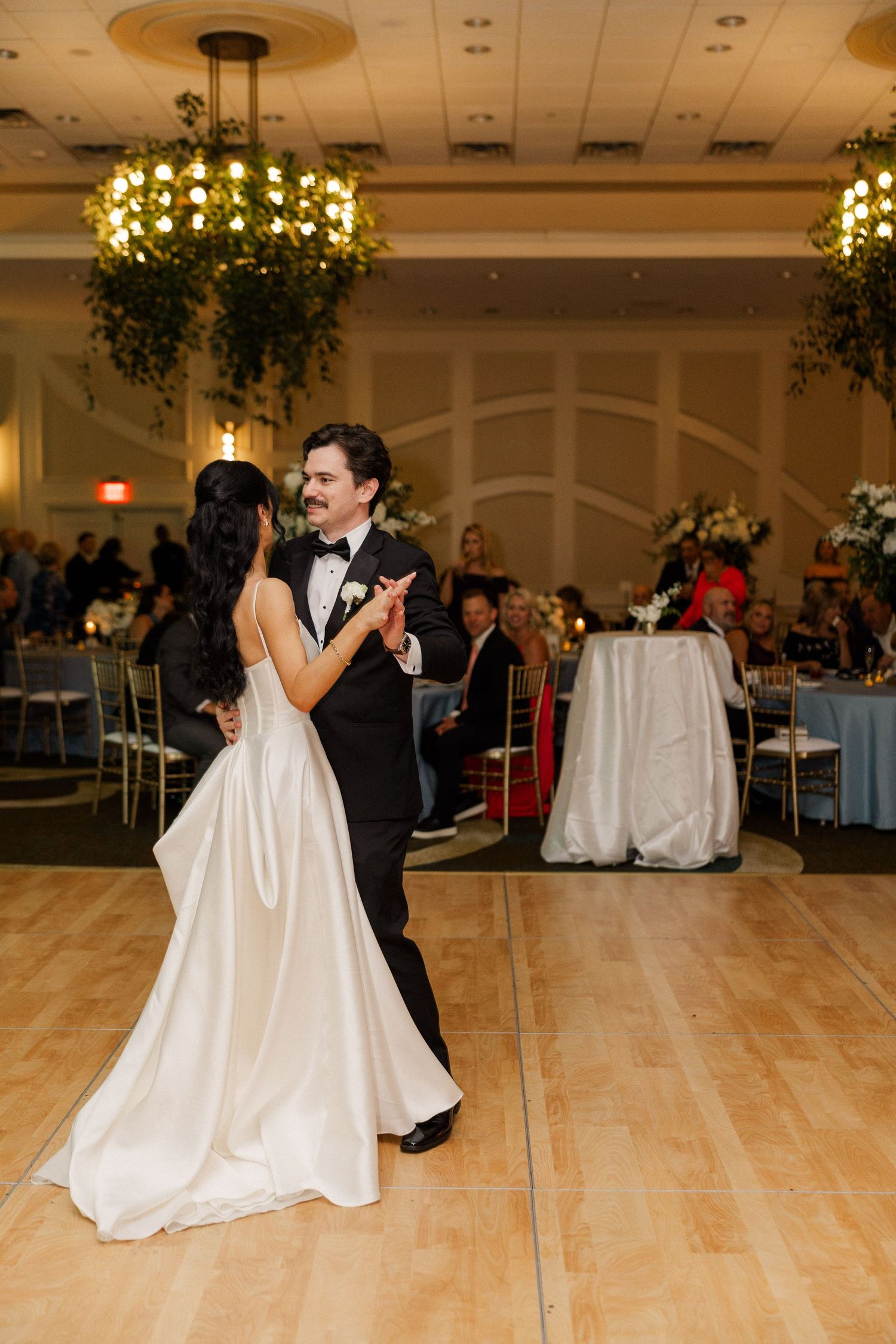Newlyweds share a joyful first dance in a decorated ballroom with guests watching nearby.