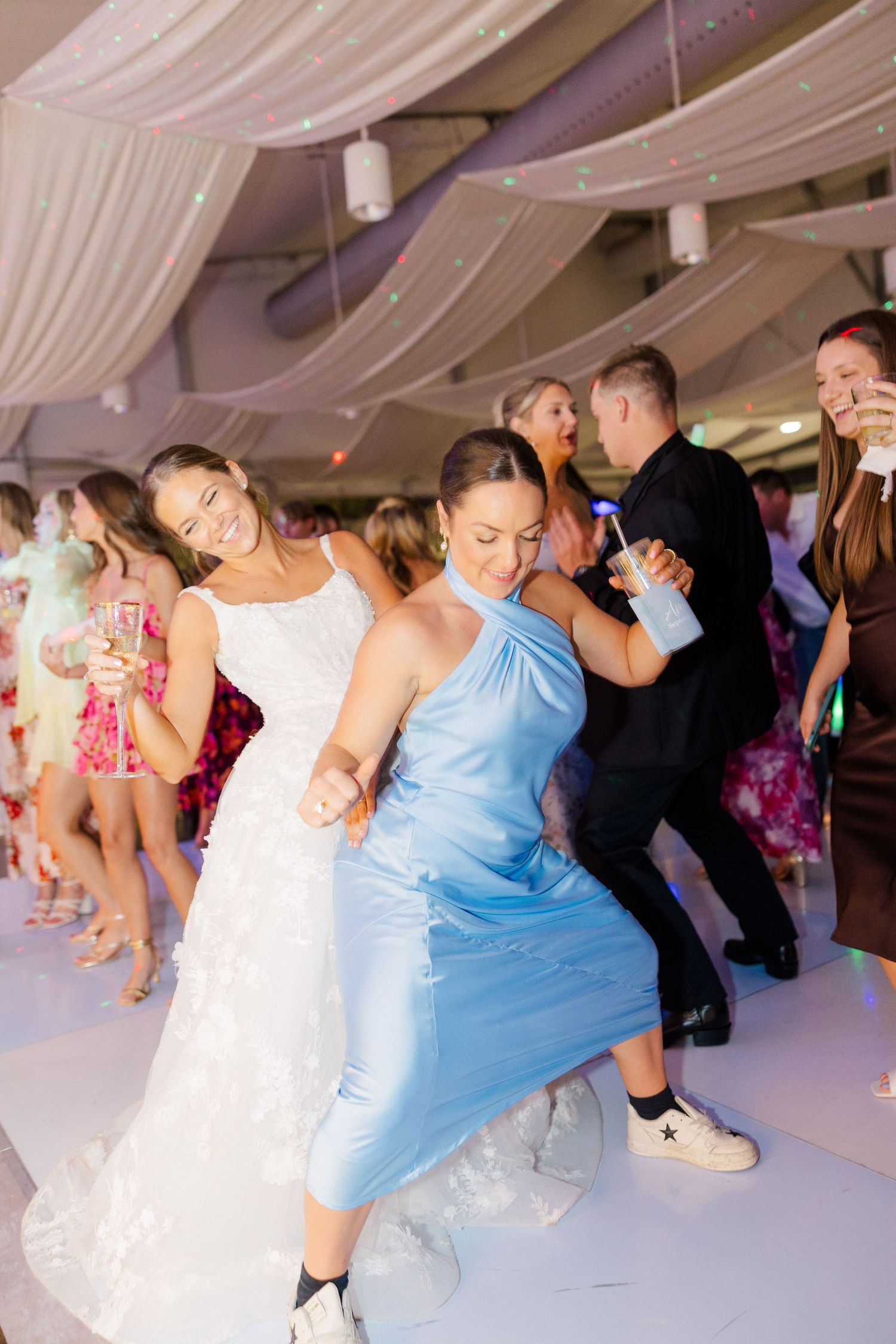 Wedding guests dance and celebrate on a white dance floor under draped ceiling decorations at a reception venue.