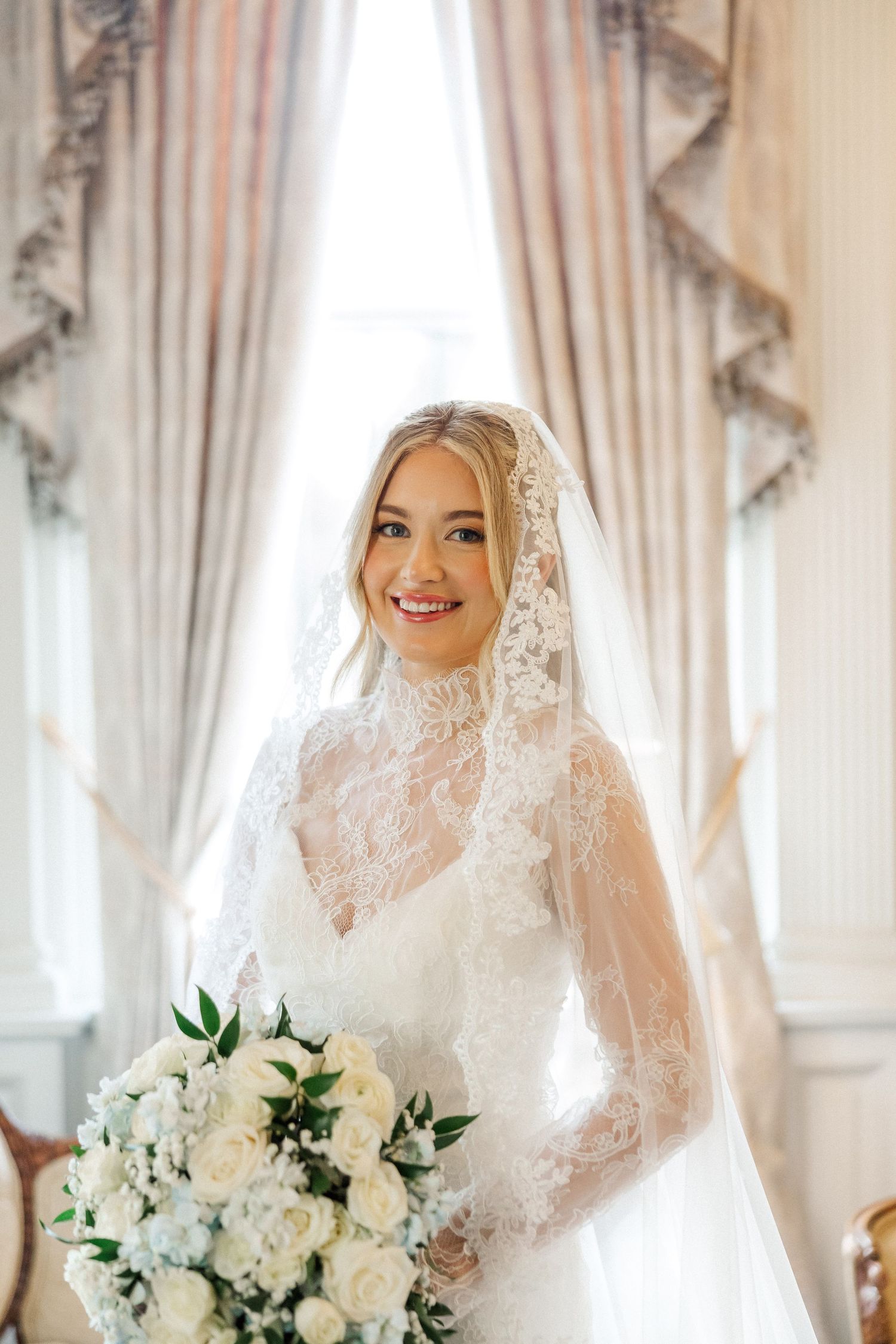 Bride holding white rose bouquet in lace veil against elegant curtained backdrop.