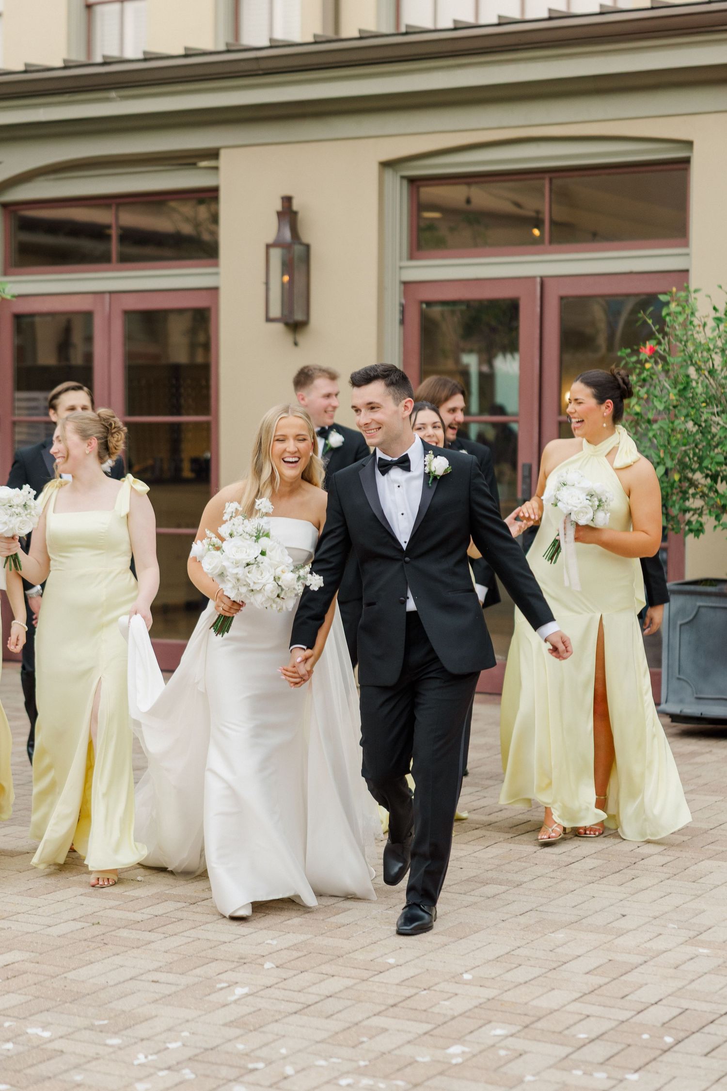 Wedding party in formal attire with bridesmaids in pale yellow dresses walking and laughing together outdoors.