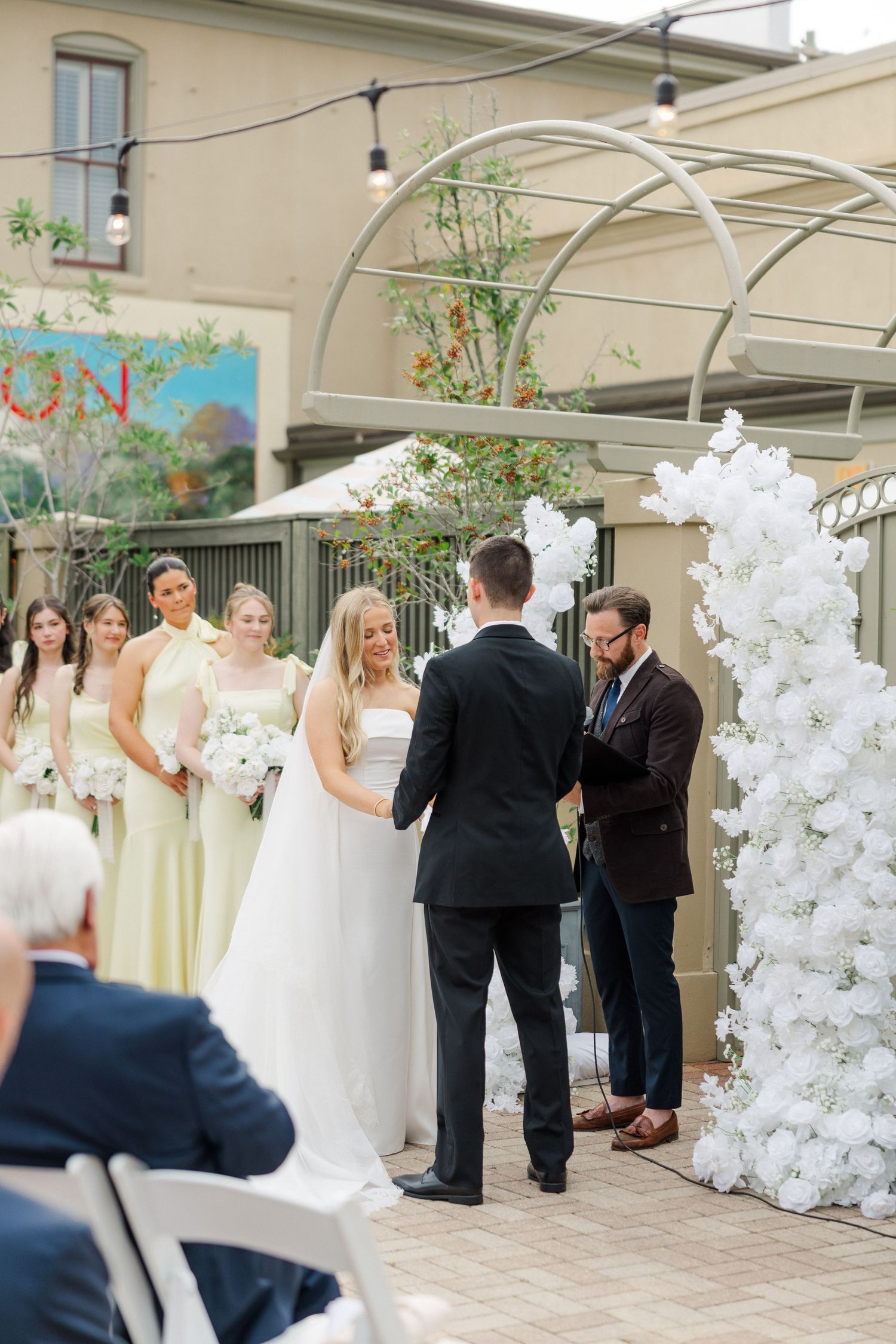 Wedding ceremony setup with white floral arrangements and guests gathering in an indoor venue.