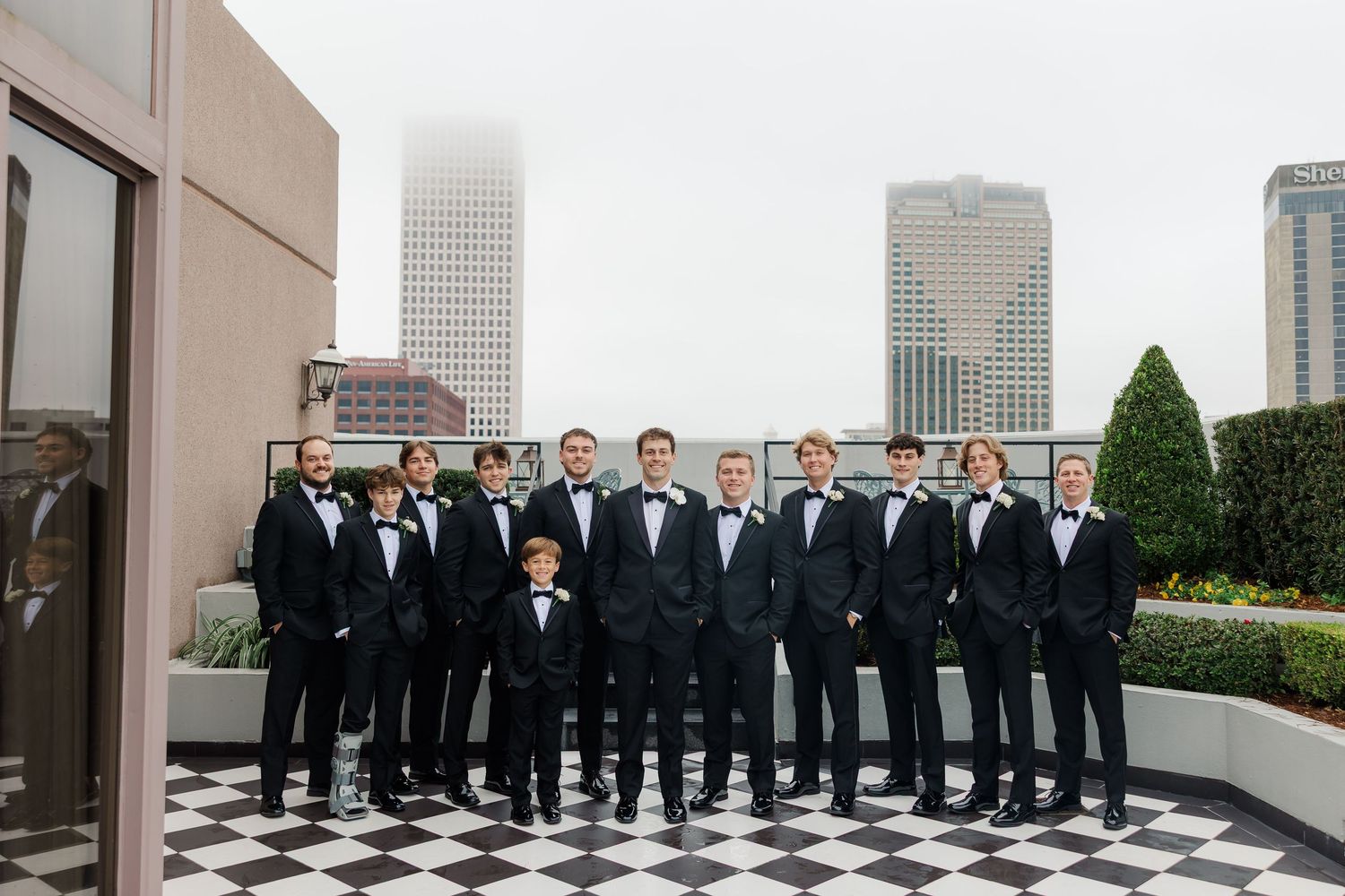 A wedding party in black tuxedos stands in a row on a rooftop with city skyscrapers visible in the background.