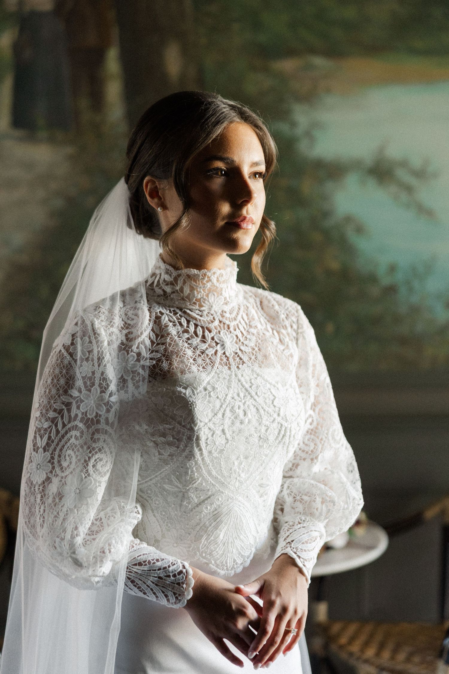 Bride in elegant high-neck lace wedding gown with balloon sleeves and flowing veil poses by window in soft light.