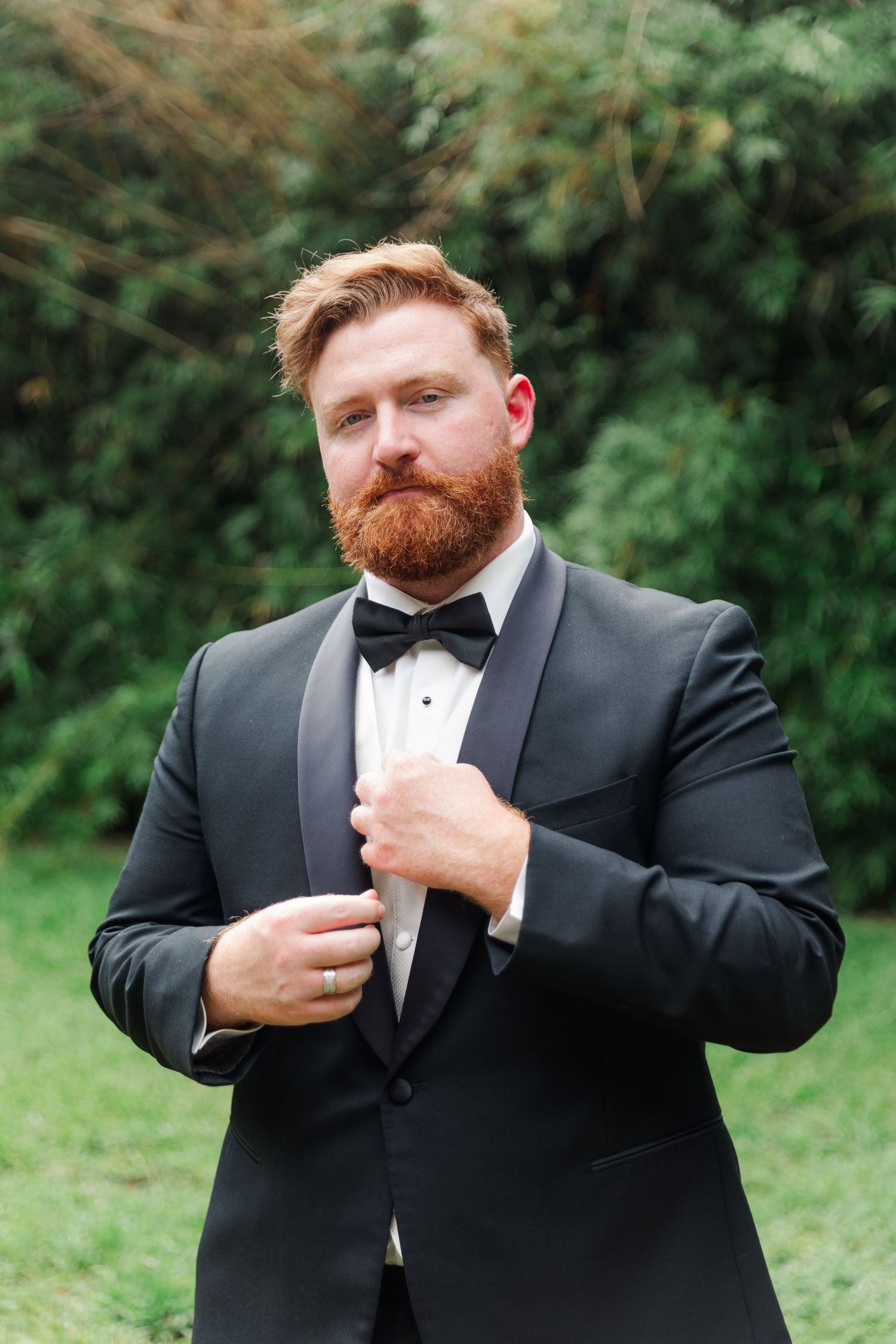 A groom in a black tuxedo and bow tie adjusts his jacket outdoors against a green background.