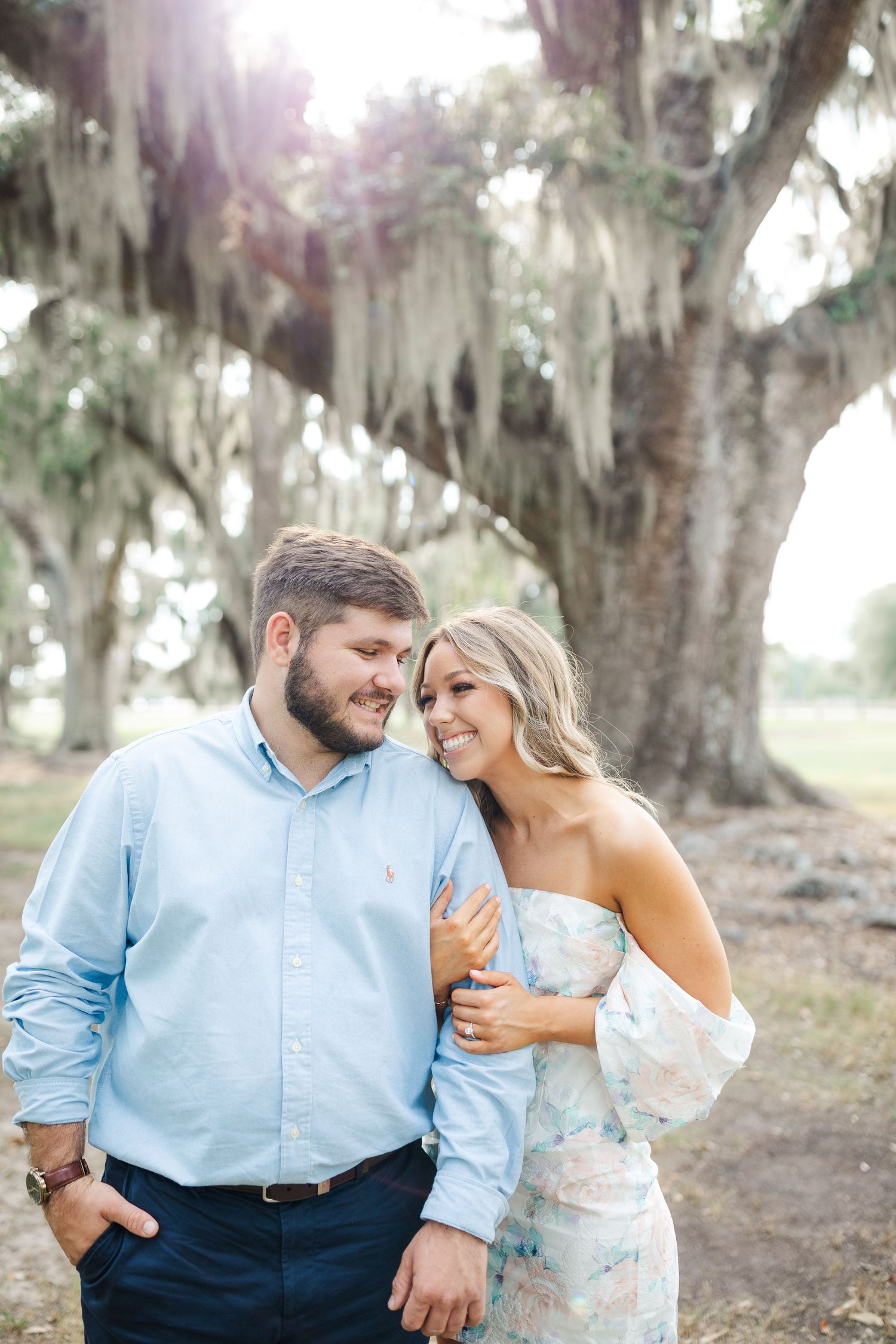 A romantic couple shares a tender moment under Spanish moss draped oak trees in a dreamy outdoor setting.