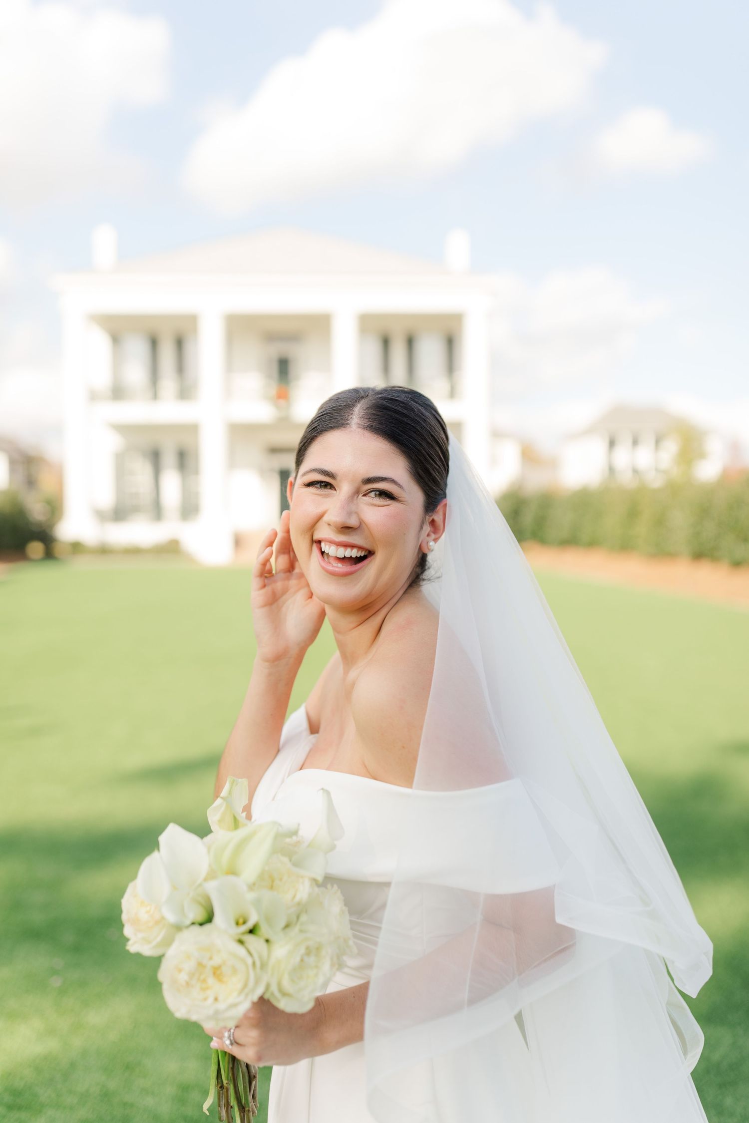 A joyful bride in a white wedding dress holding a bouquet of white roses laughs on a sunny lawn with a mansion behind her.