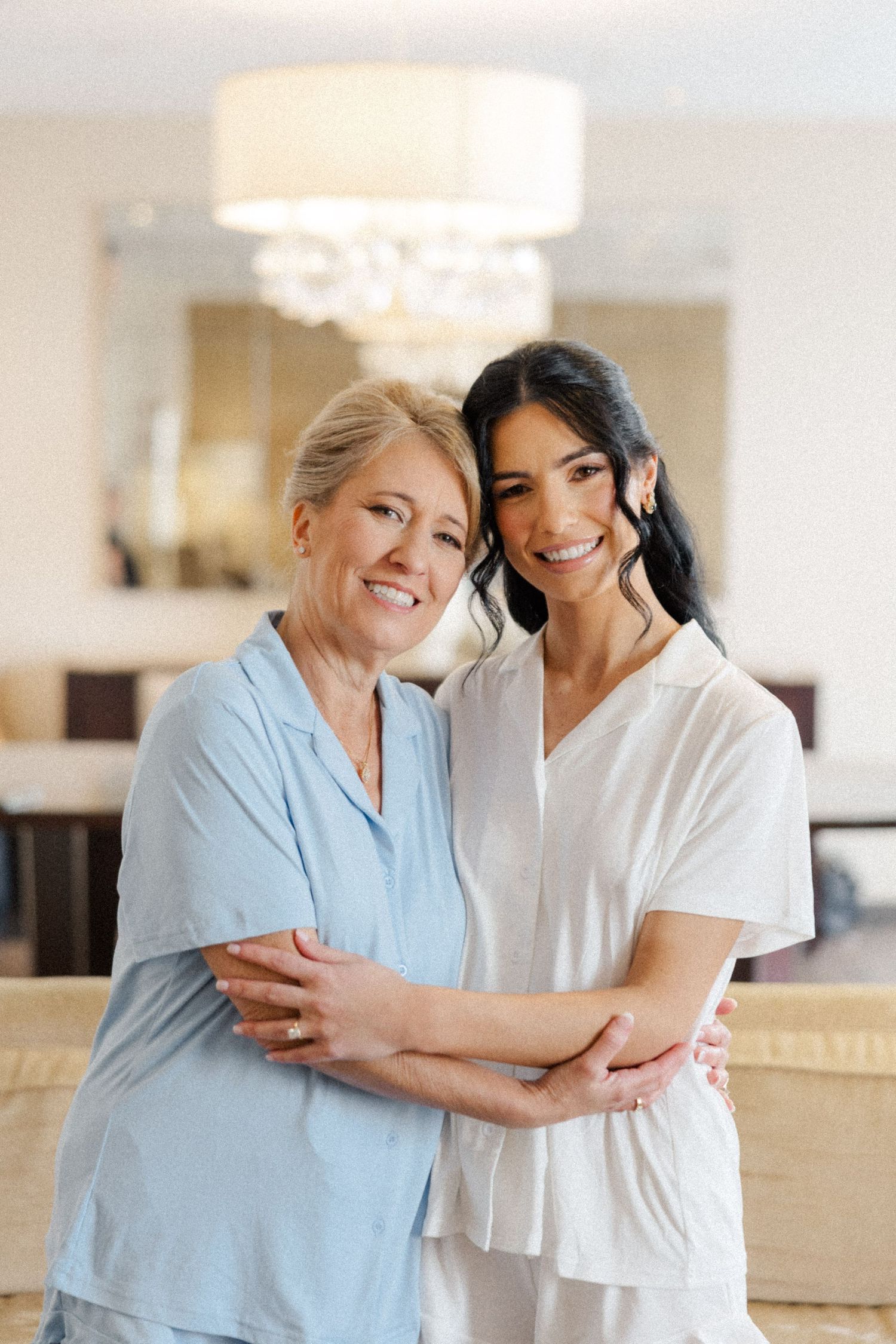 Two women in pastel tops share a warm embrace and smile in an elegant hotel suite.
