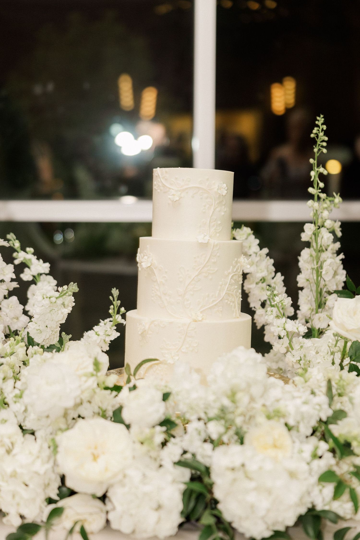 Elegant four-tier white wedding cake surrounded by white peonies and floral arrangements.