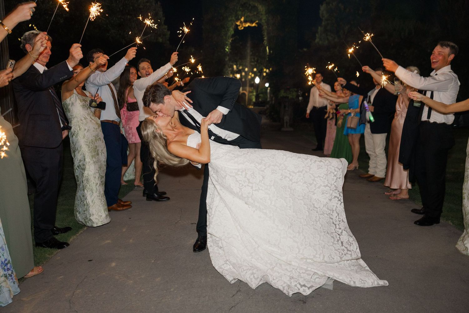 Groom dips bride dramatically during sparkler send-off exit at nighttime wedding celebration.