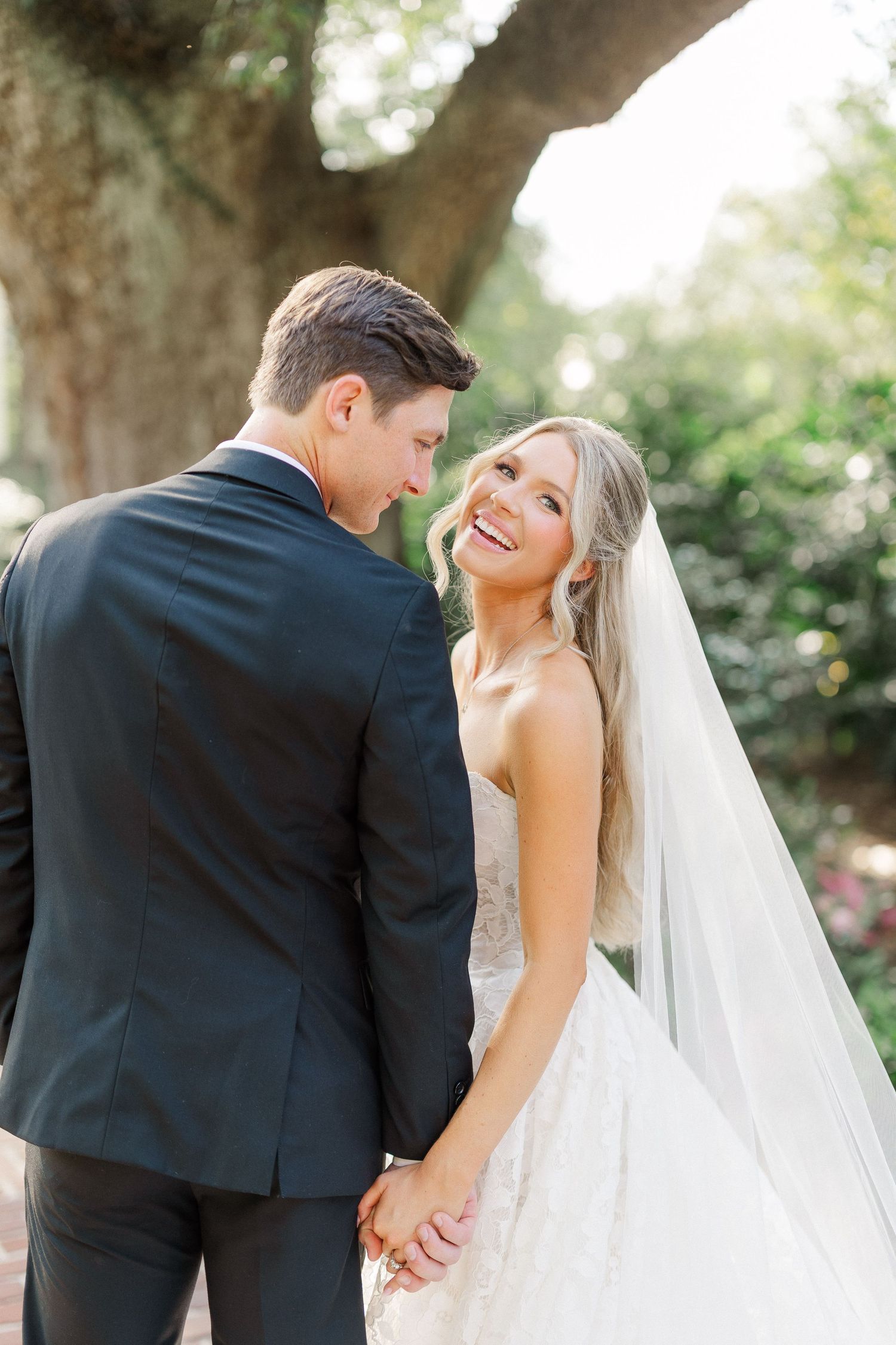 Bride in lace gown and veil smiles over shoulder while holding hands with groom in black suit outdoors.