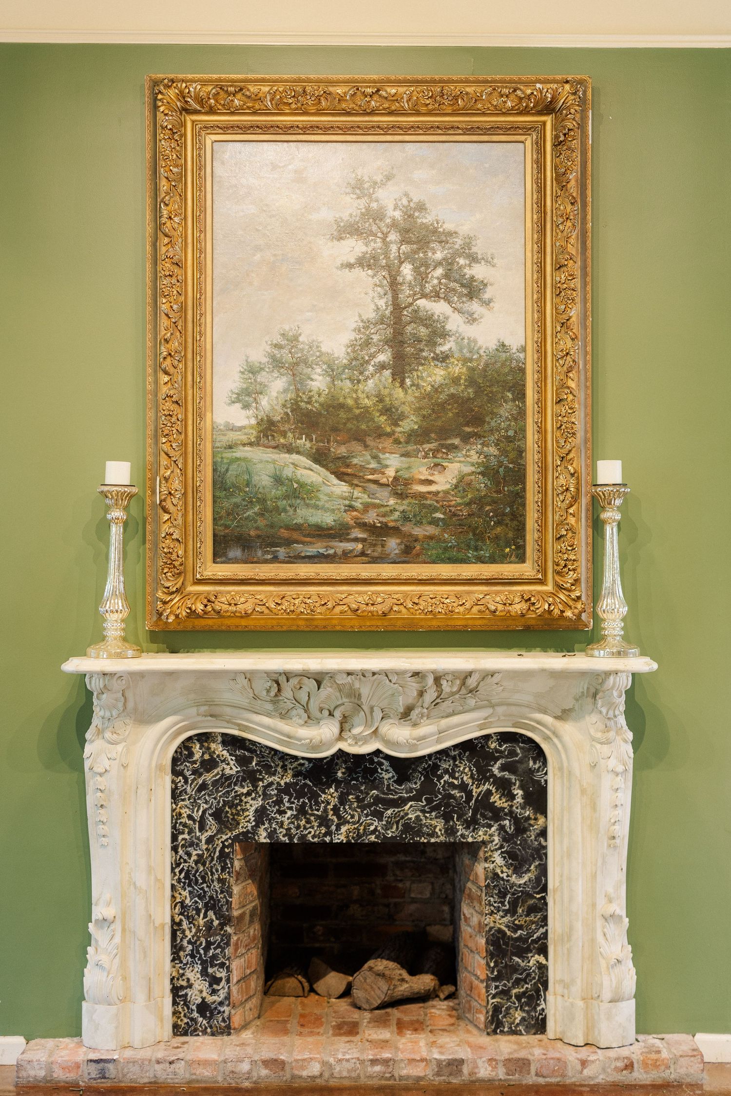 Elegant marble fireplace with ornate gold-framed landscape painting above mantel, flanked by candlesticks on green wall.