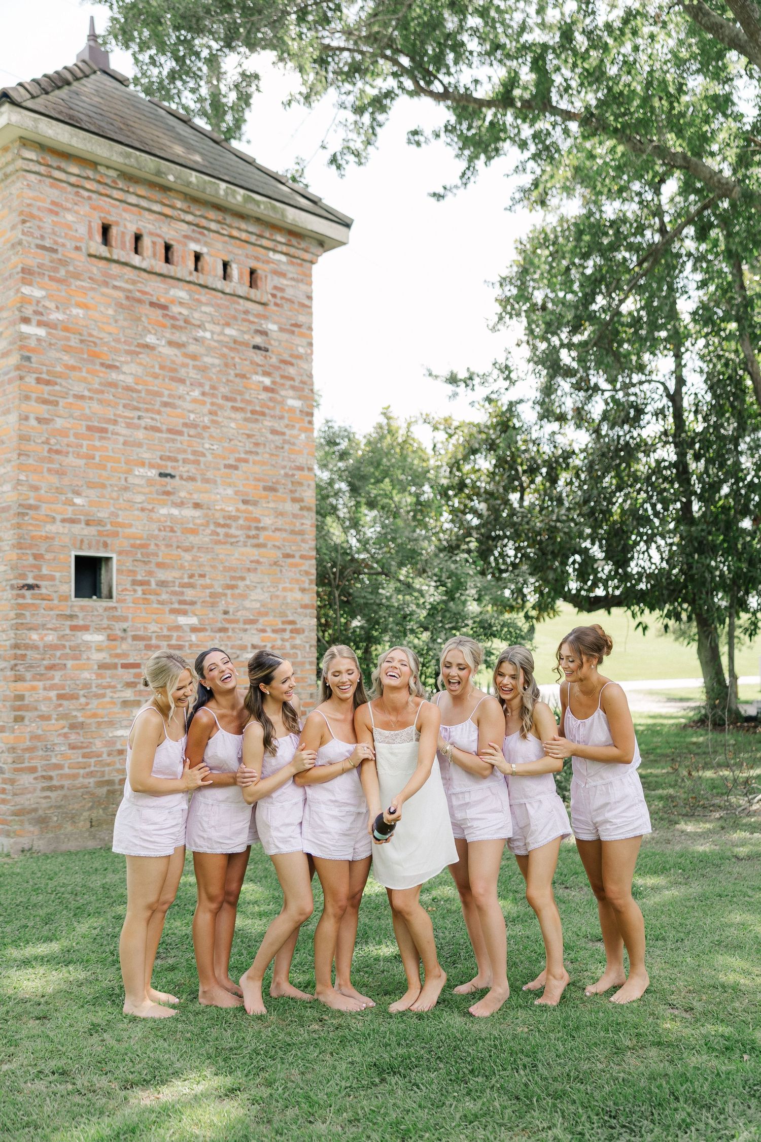 Bridesmaids in pink satin sets celebrate with bride in white, popping champagne beside a brick tower on green lawn.