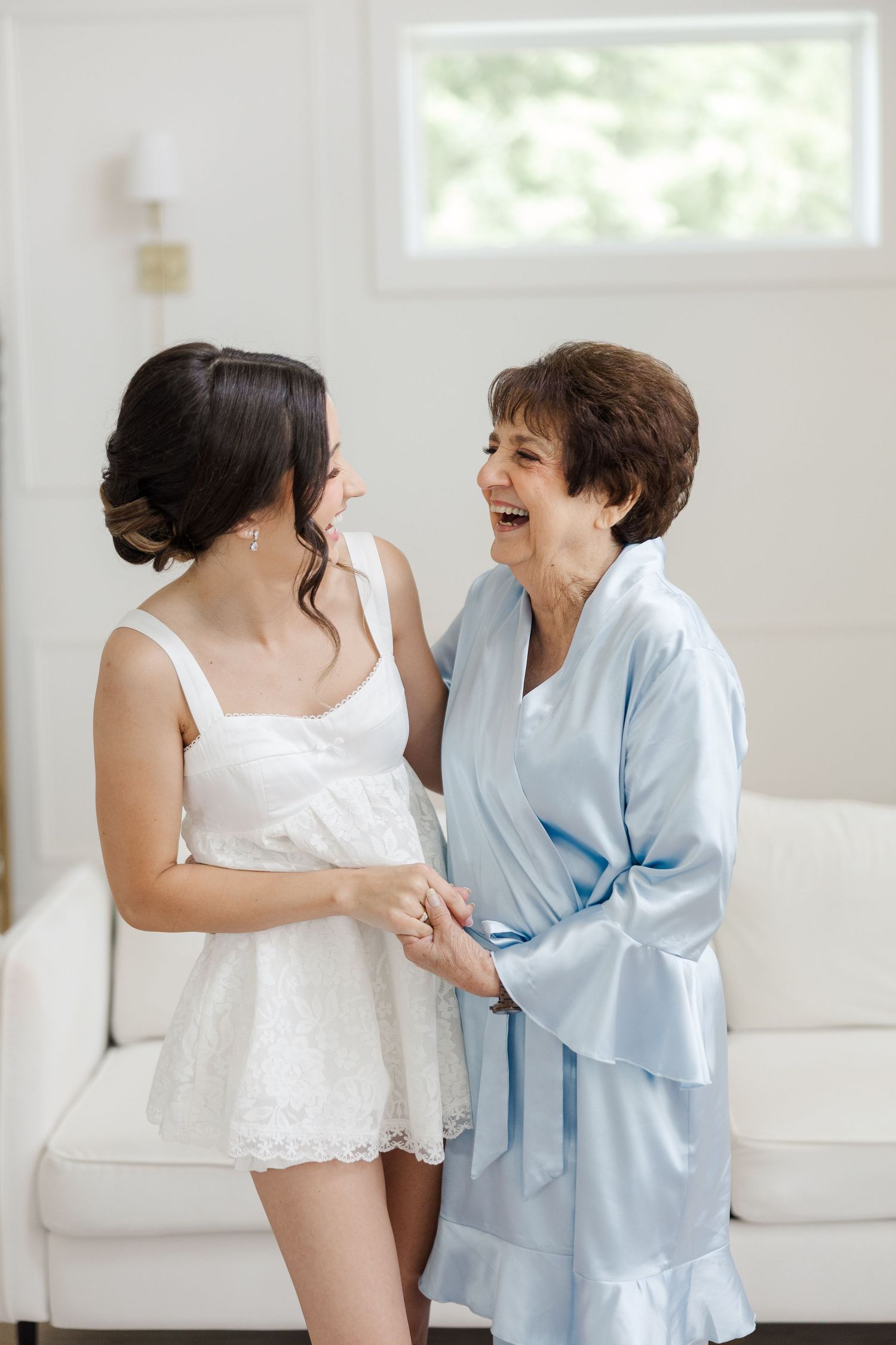 Bride and older woman laughing together in white bridal suite during wedding morning preparations.