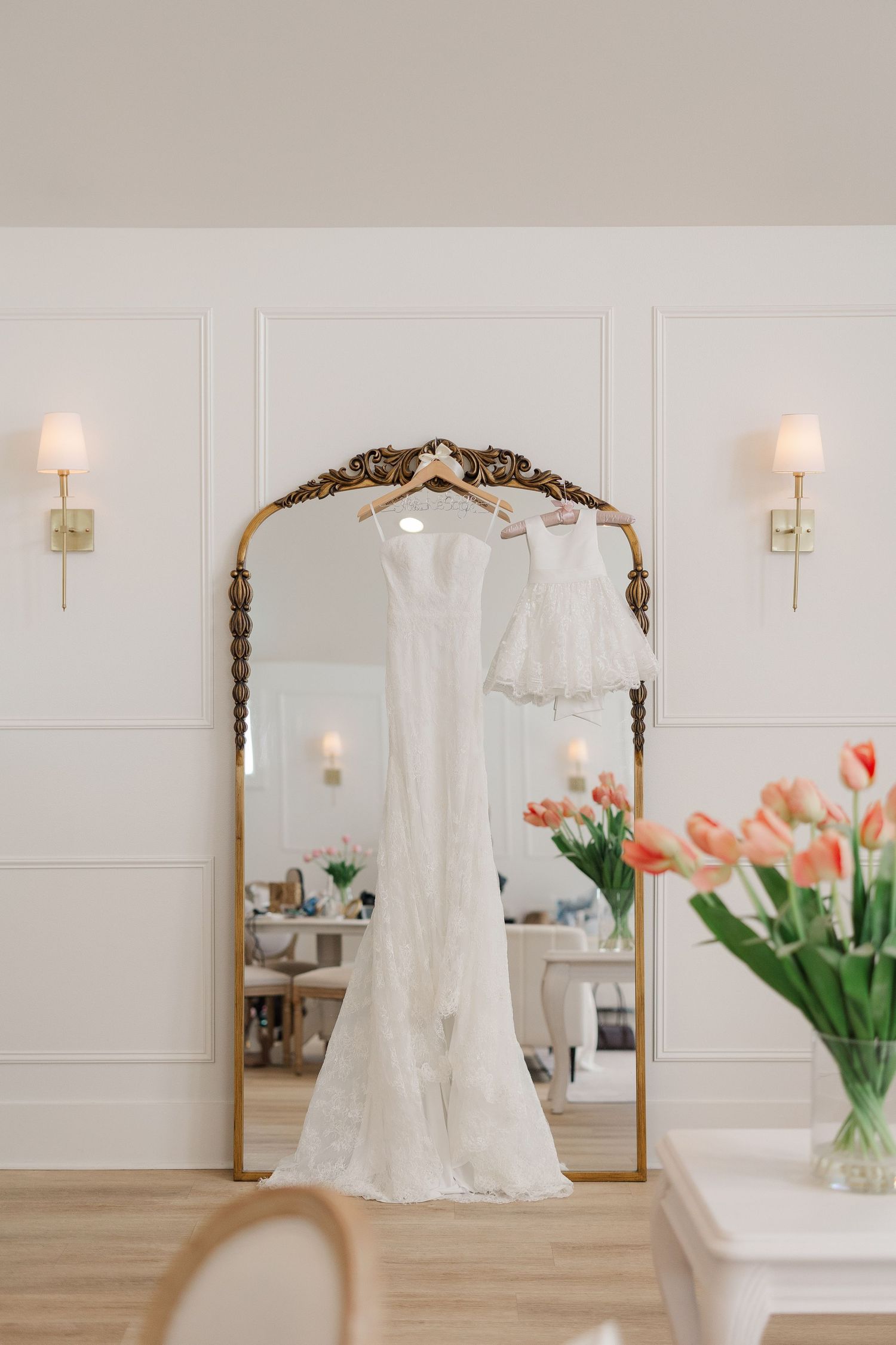 Elegant white lace wedding gown and flower girl dress hanging on an ornate gold mirror in a bright, minimalist bridal suite.