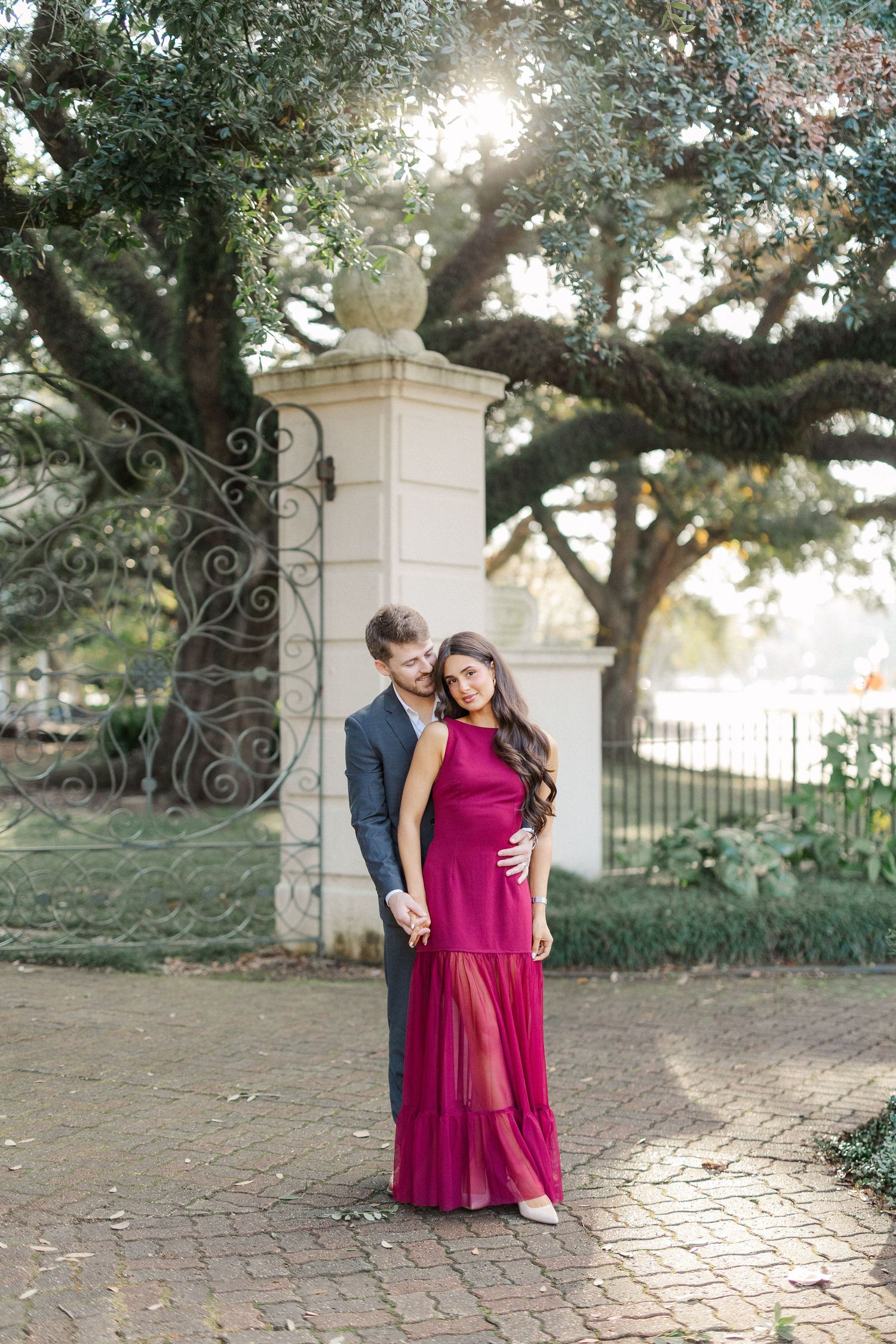 A couple poses together in a romantic outdoor setting with stone pillars and trees in the background.