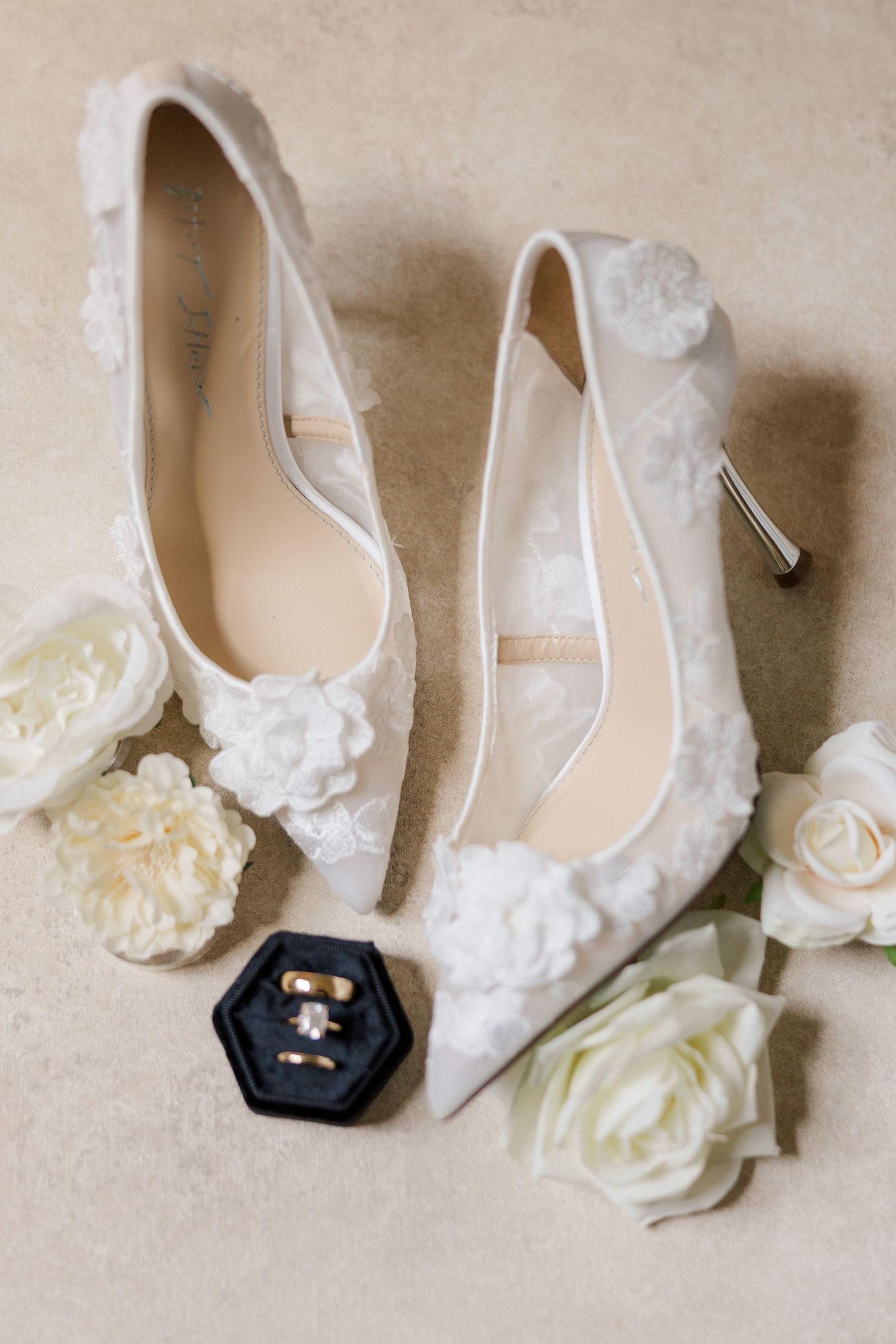 Elegant white lace bridal shoes with delicate floral details and a small black decorative accent piece displayed in a row.