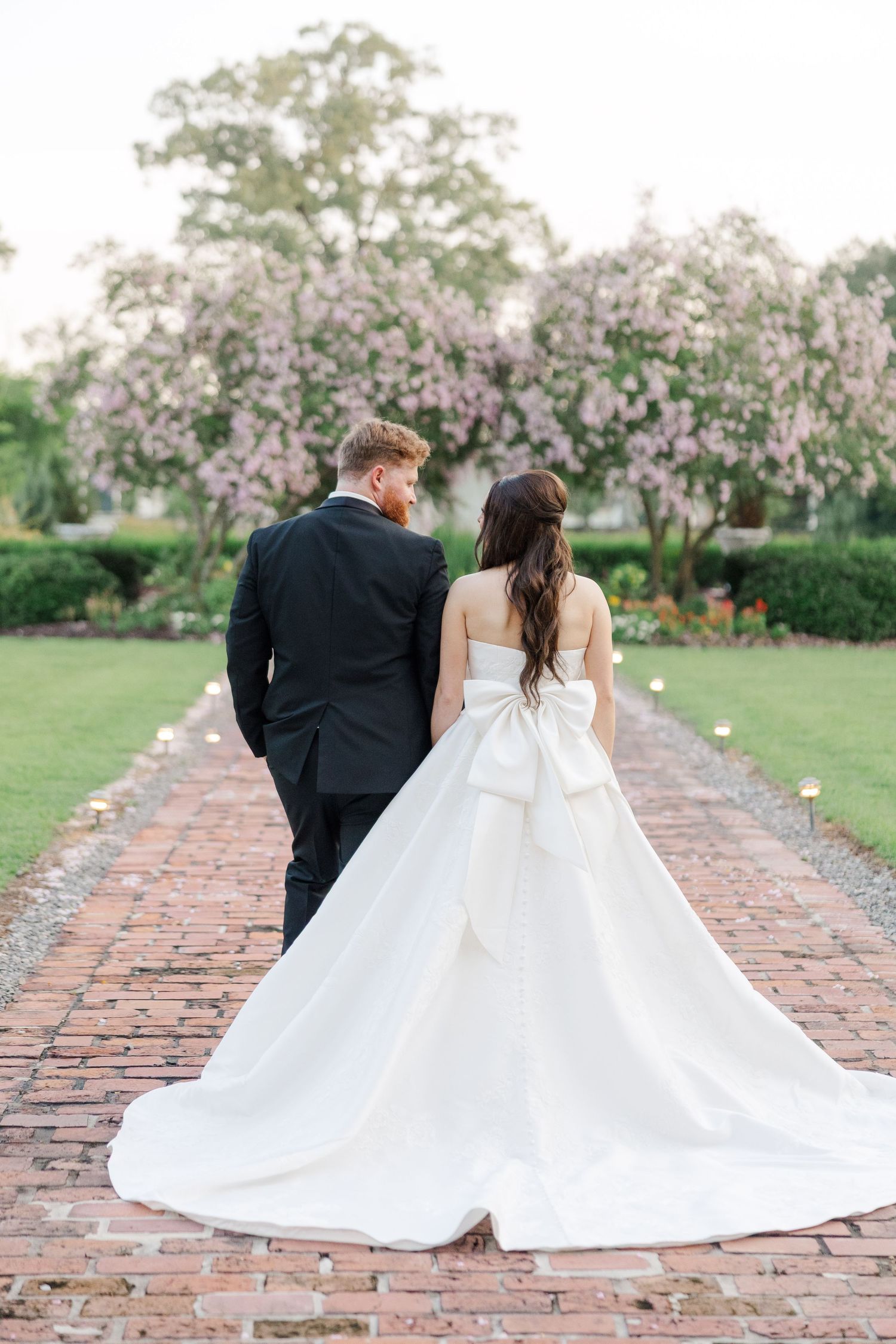 A couple in wedding attire sits together on a brick path surrounded by blooming cherry blossom trees.
