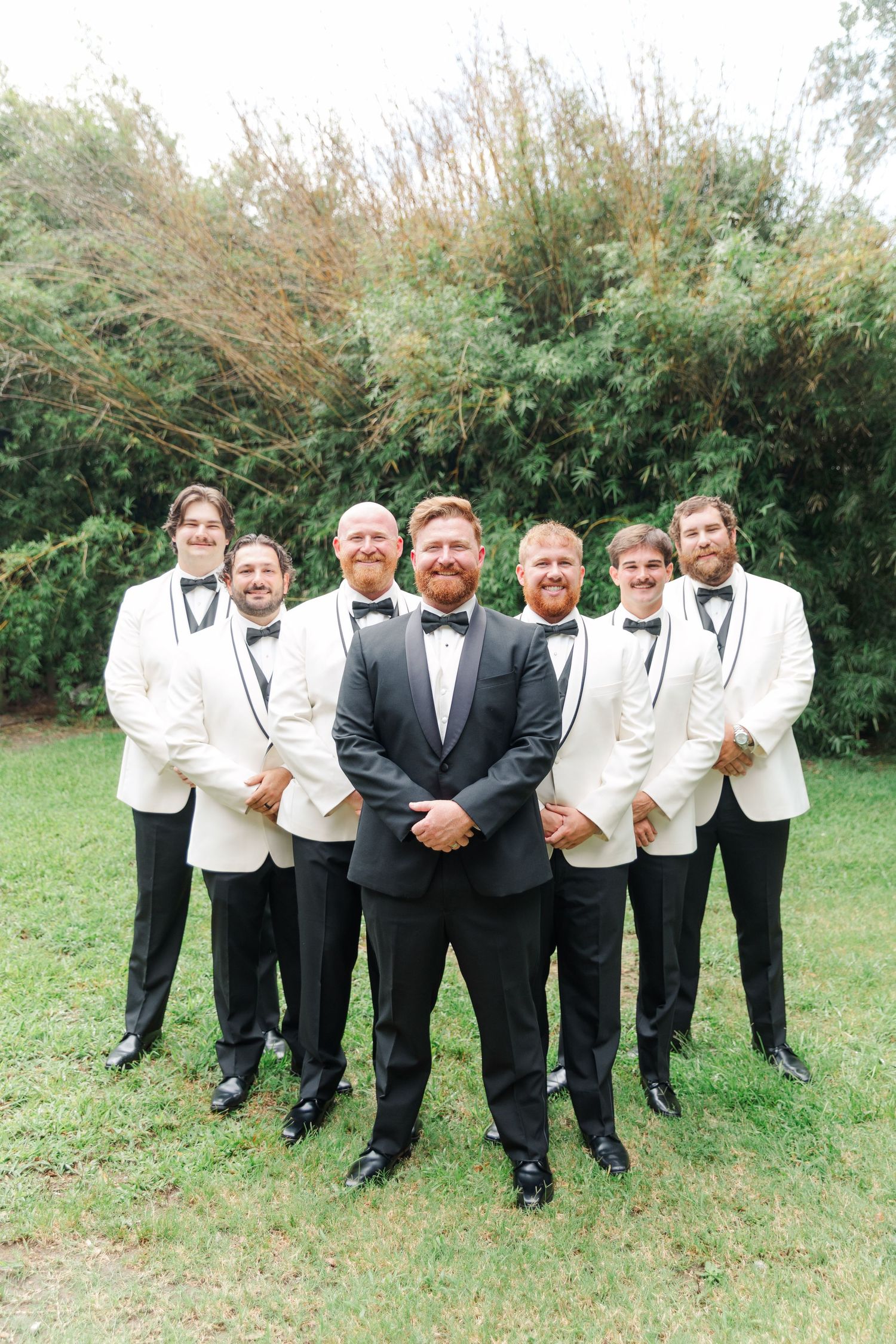 Wedding party in black and white tuxedos posing outdoors on a grassy lawn.
