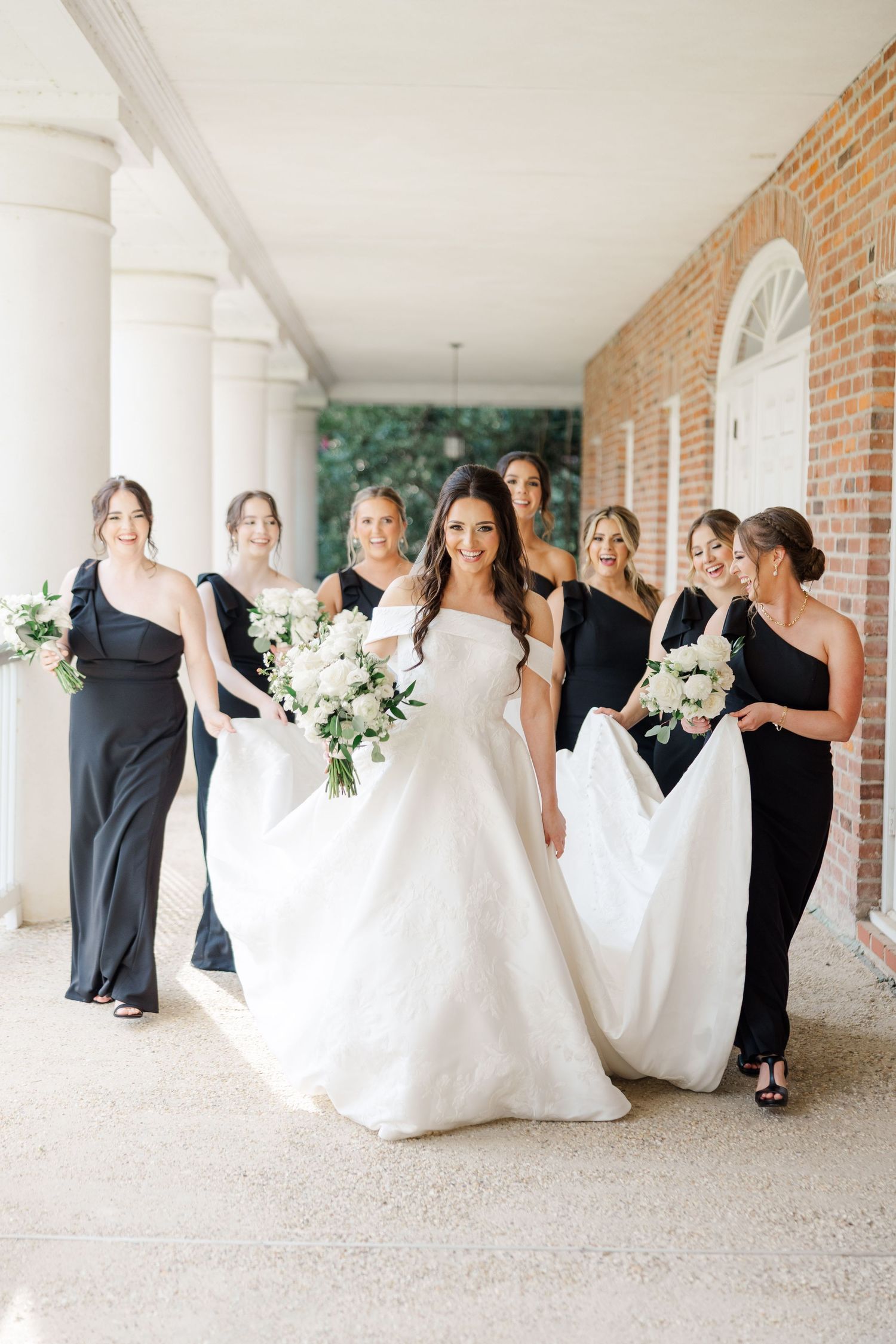 Wedding party poses in elegant black dresses and white gowns along a columned walkway.
