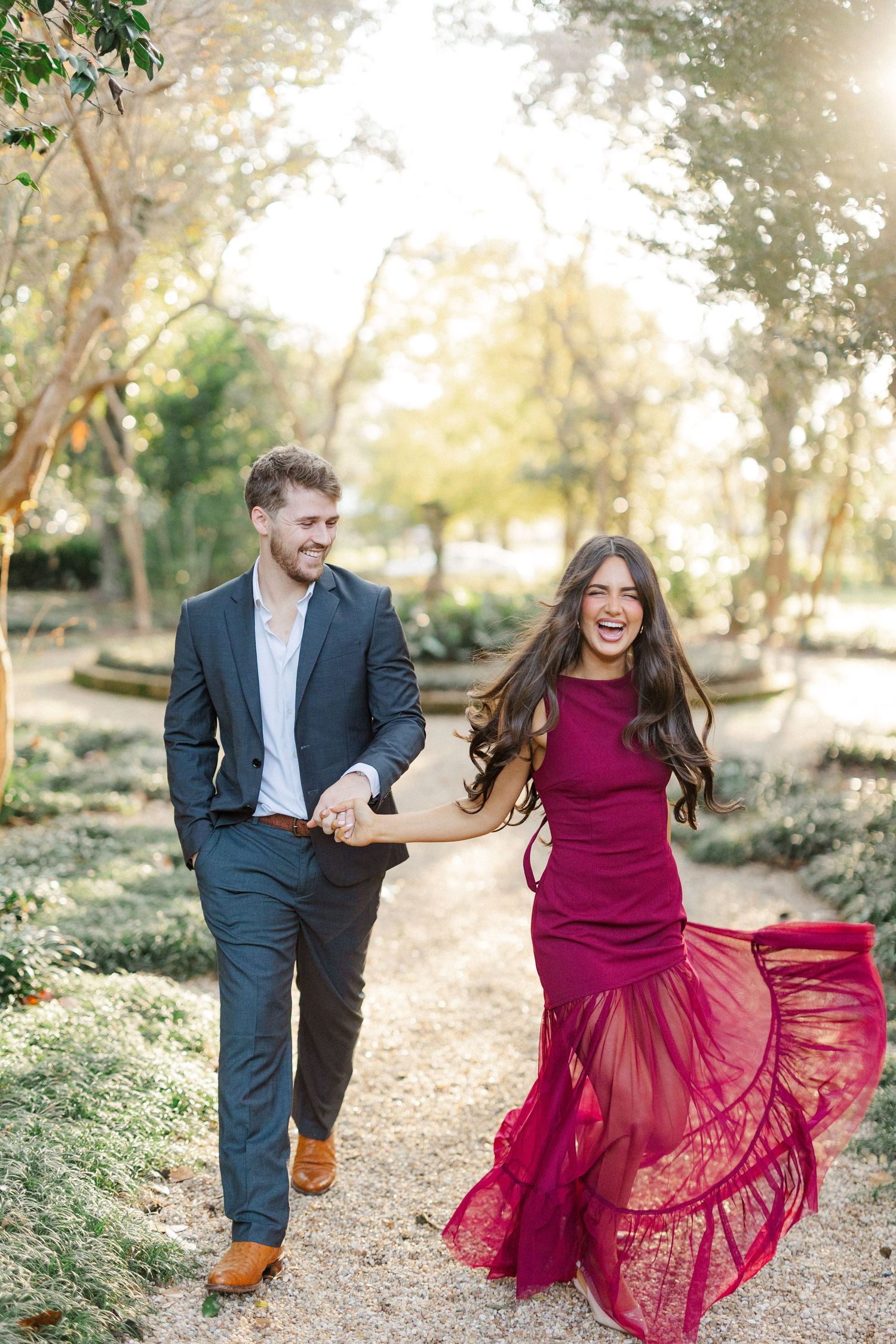 A couple walks hand in hand on a garden path as her flowing burgundy dress catches the breeze.