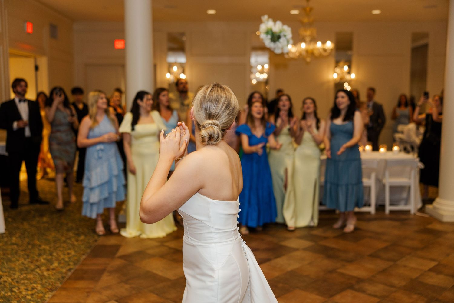 Wedding reception in a grand ballroom with white columns and chandeliers as guests gather around the dance floor.