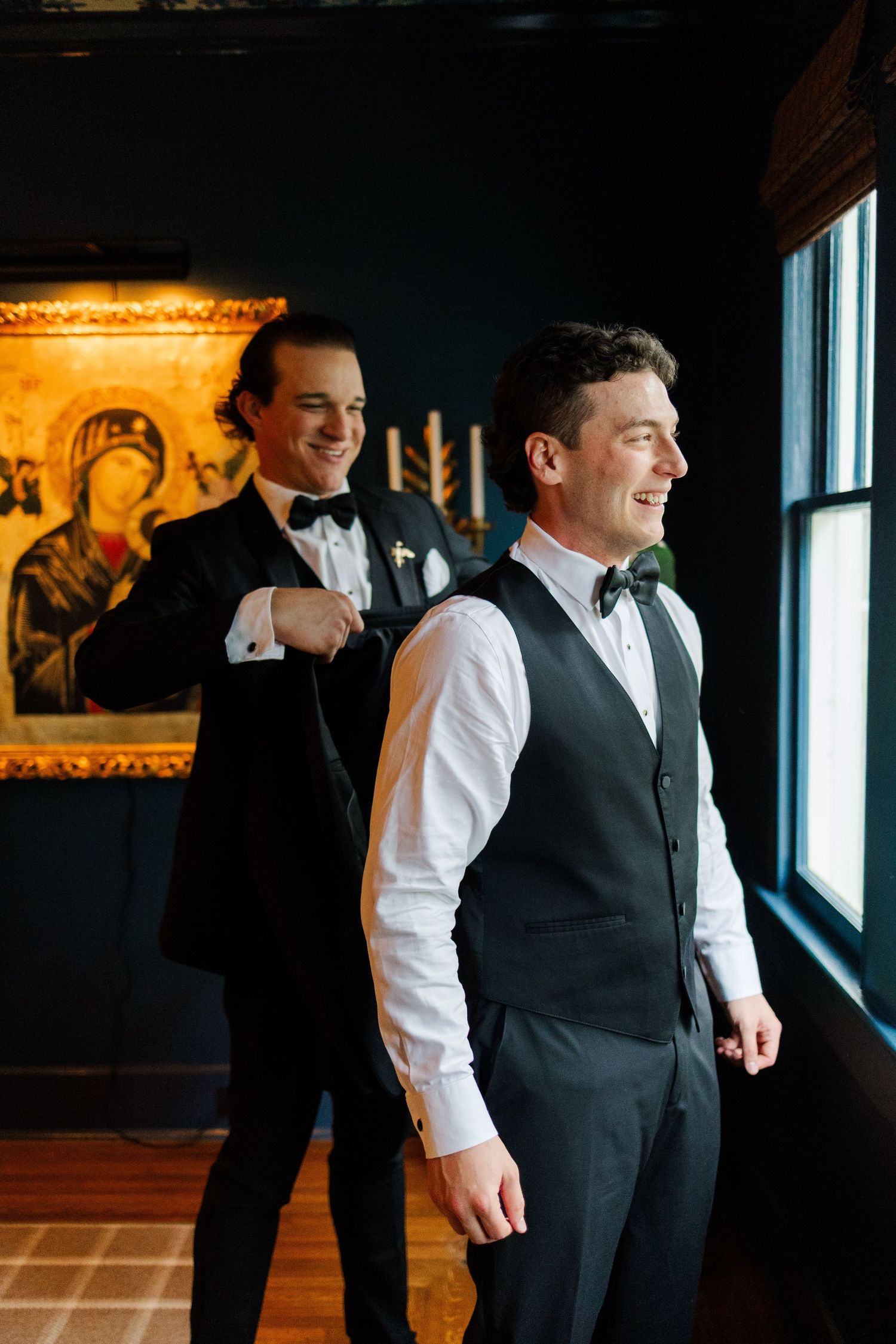 Two people in formal attire stand by large windows in a dark room with warm lighting and artwork on the walls.