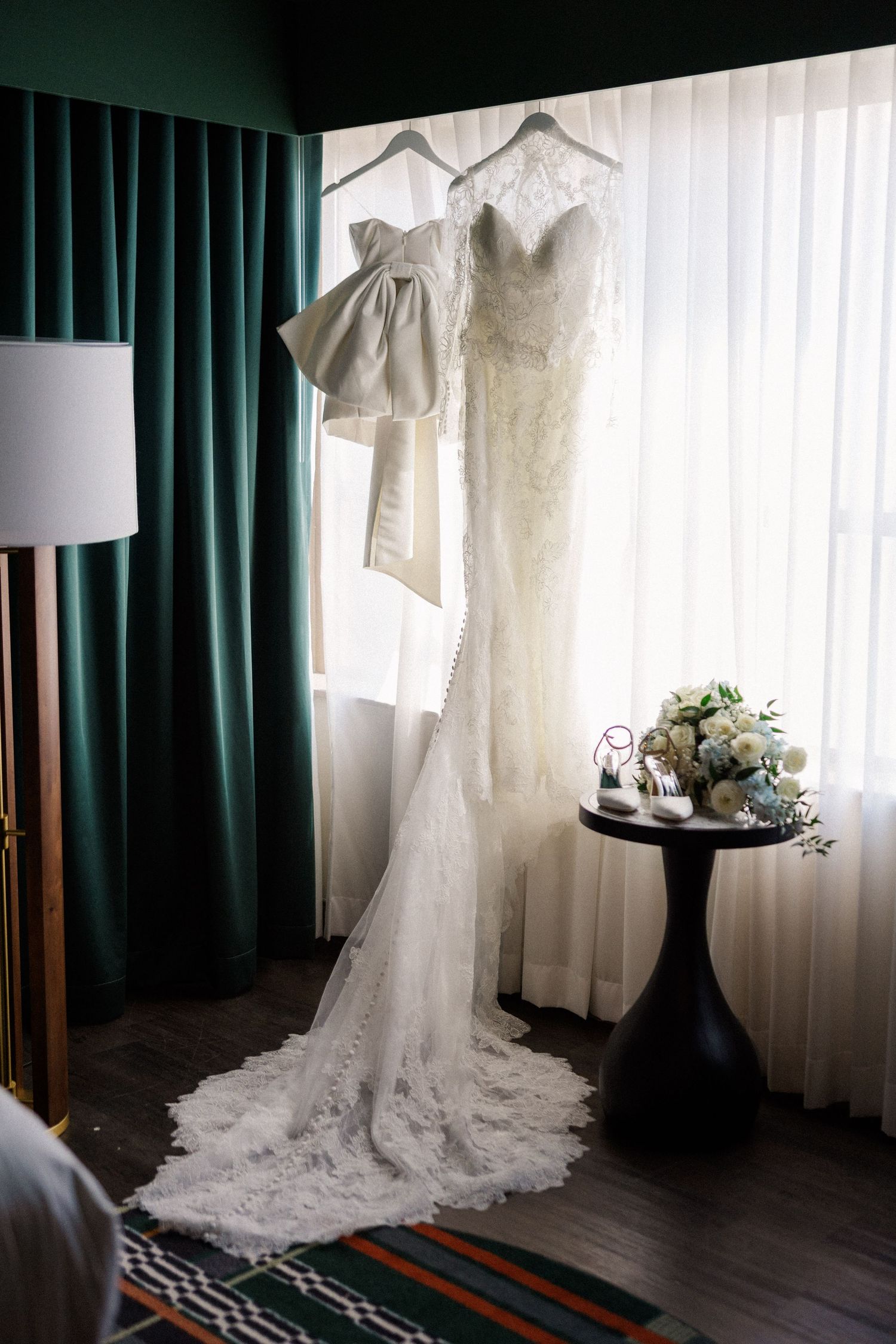 Elegant lace wedding dress hanging by window with emerald green curtains and small floral arrangement.