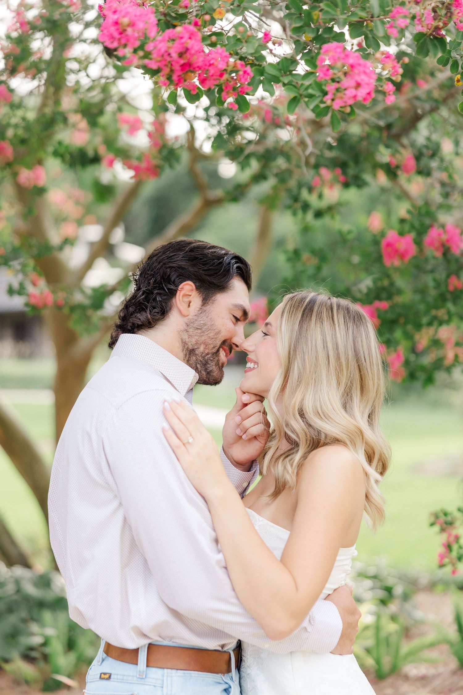 Romantic couple embraces under blooming pink crepe myrtle trees in a lush garden setting.