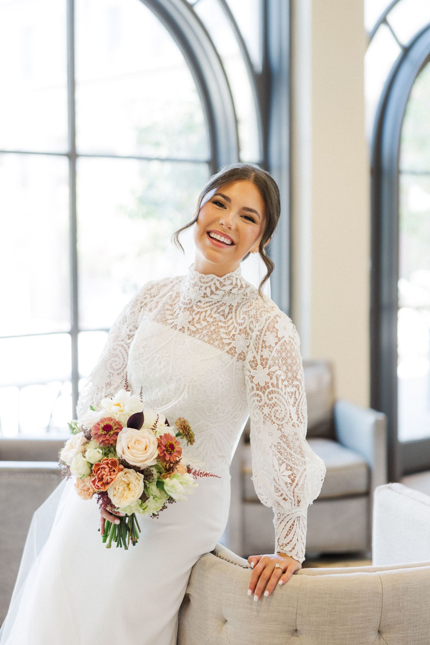 Joyful bridal portraits in a bright indoor setting showcasing a high-neck lace wedding gown and fresh floral bouquet.