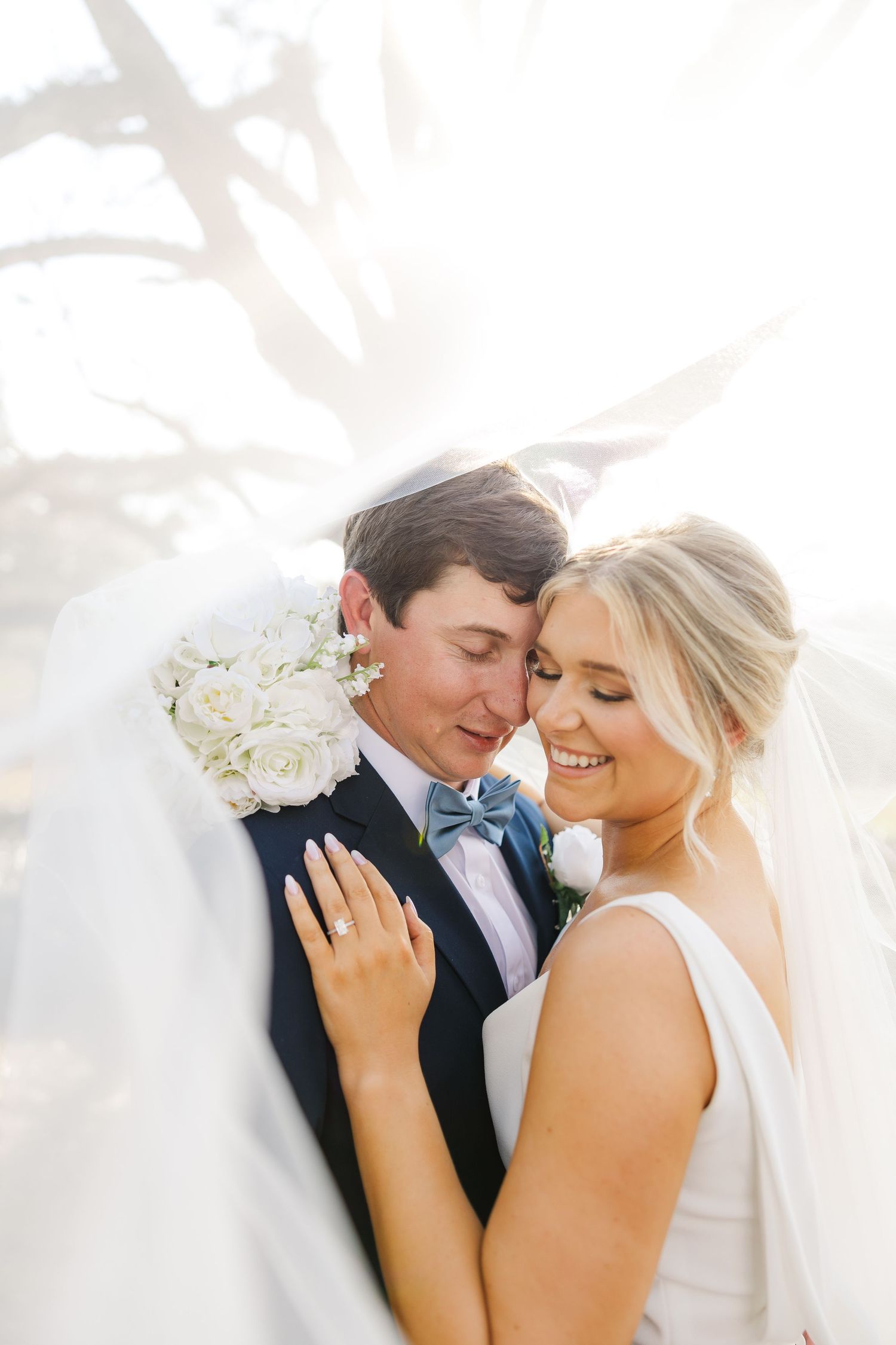 A romantic newlywed couple embraces in soft sunlight with white floral bouquet and flowing veil creating an ethereal moment.