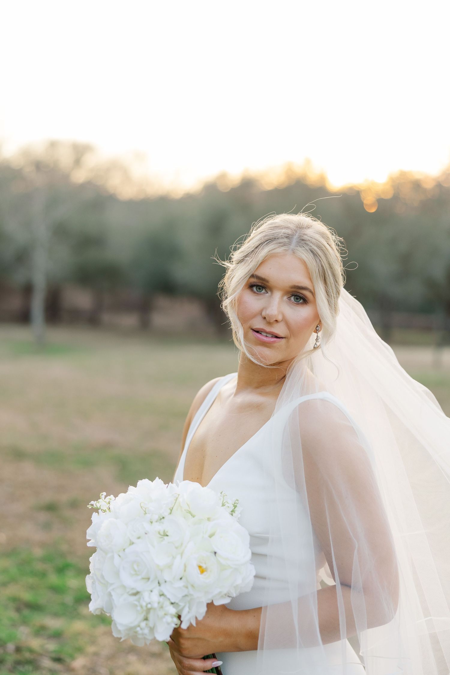 A bride in a white wedding dress holds a bouquet of white flowers during golden hour outdoors.