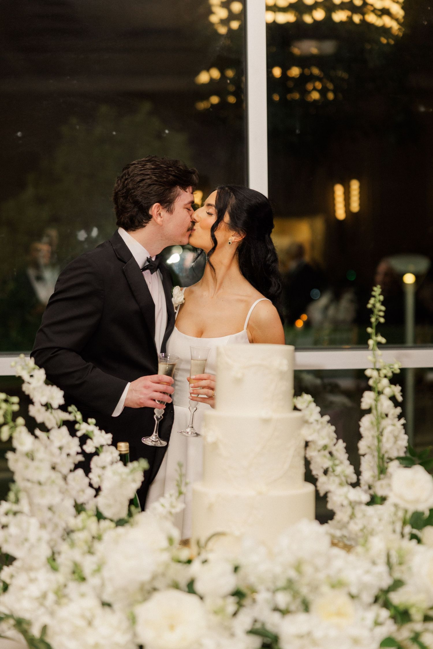 Newlyweds share a kiss holding champagne glasses beside white tiered wedding cake.