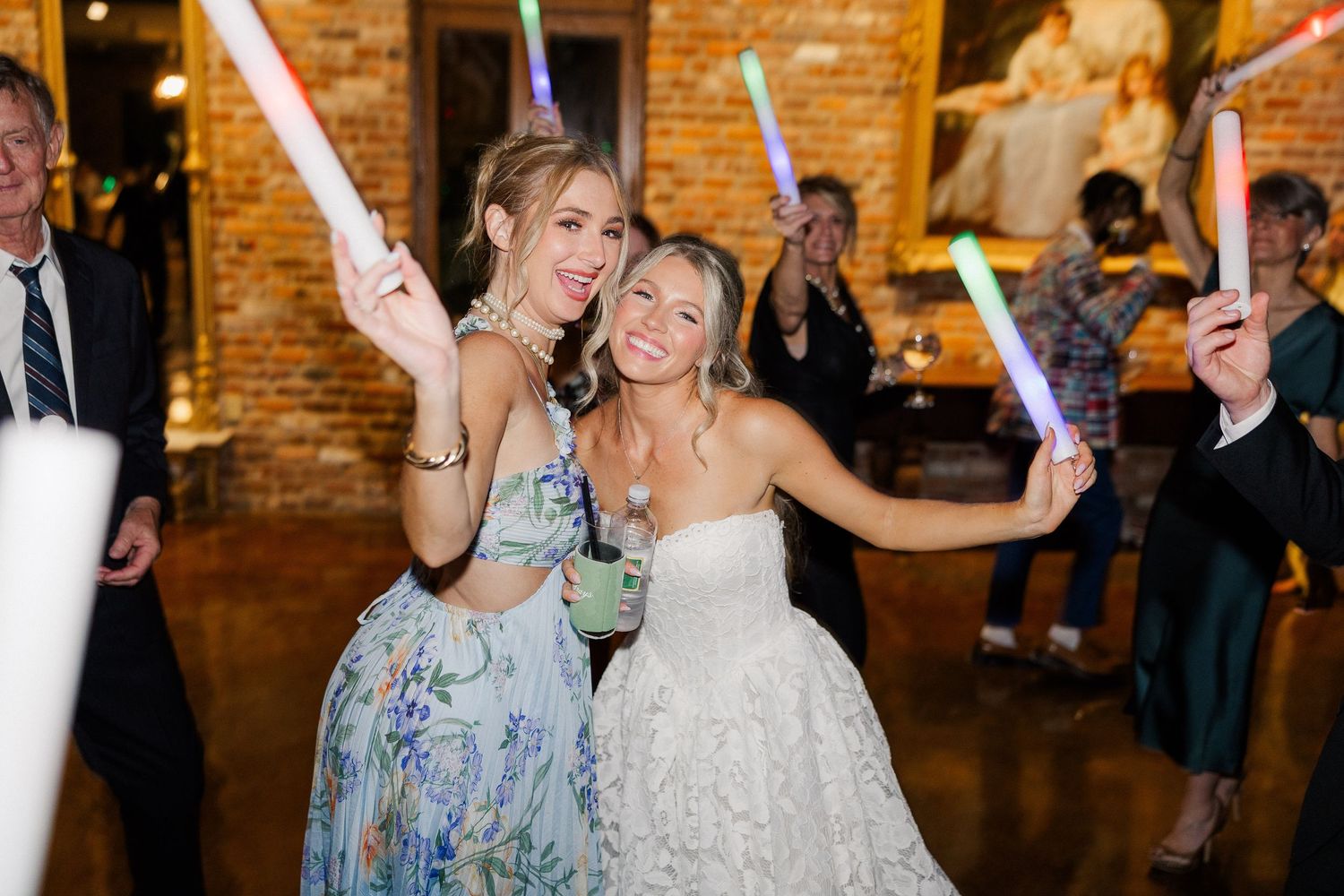 Bride in lace gown and guest in floral dress celebrate with colorful glow sticks at wedding reception.