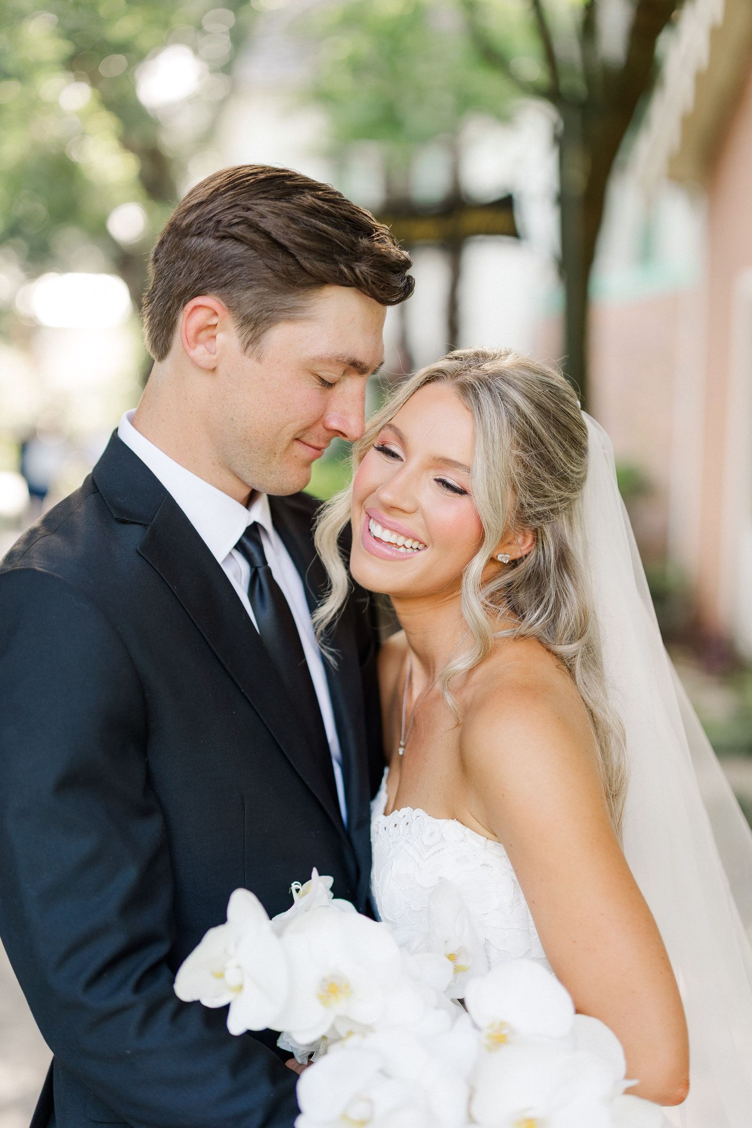 Smiling bride in white lace gown holding white orchid bouquet with groom in black tuxedo outdoors.