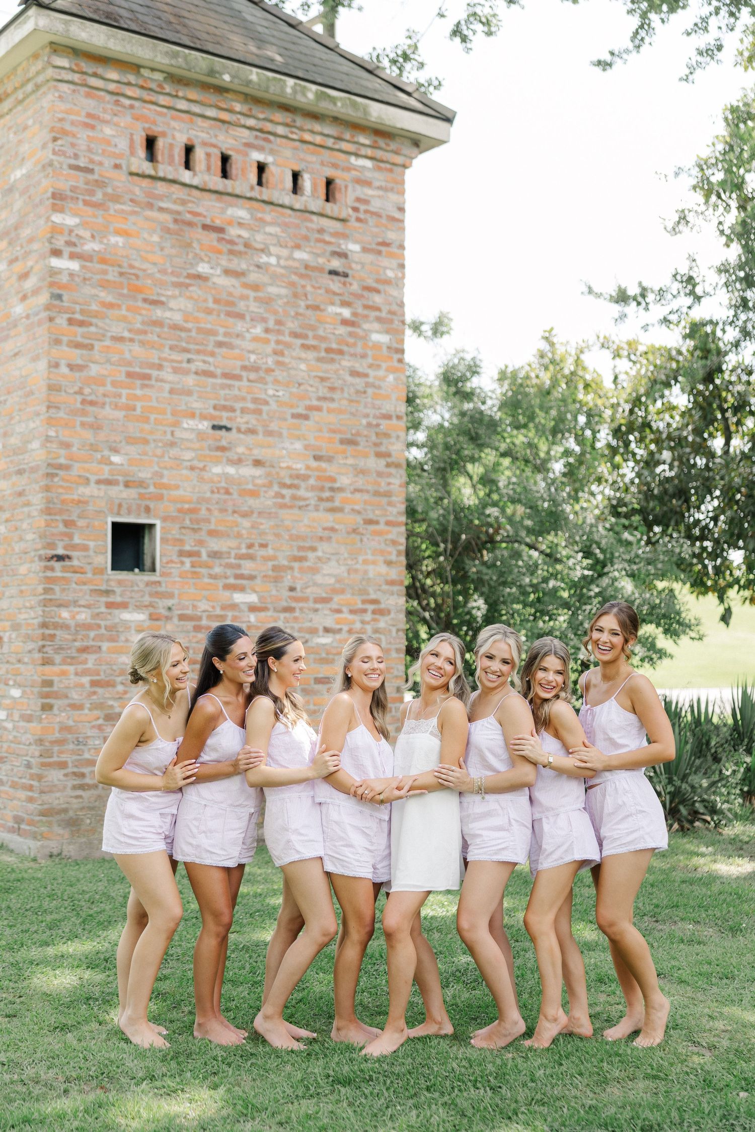 Bride and bridesmaids in matching pink pajamas posing barefoot by historic brick tower outdoors.