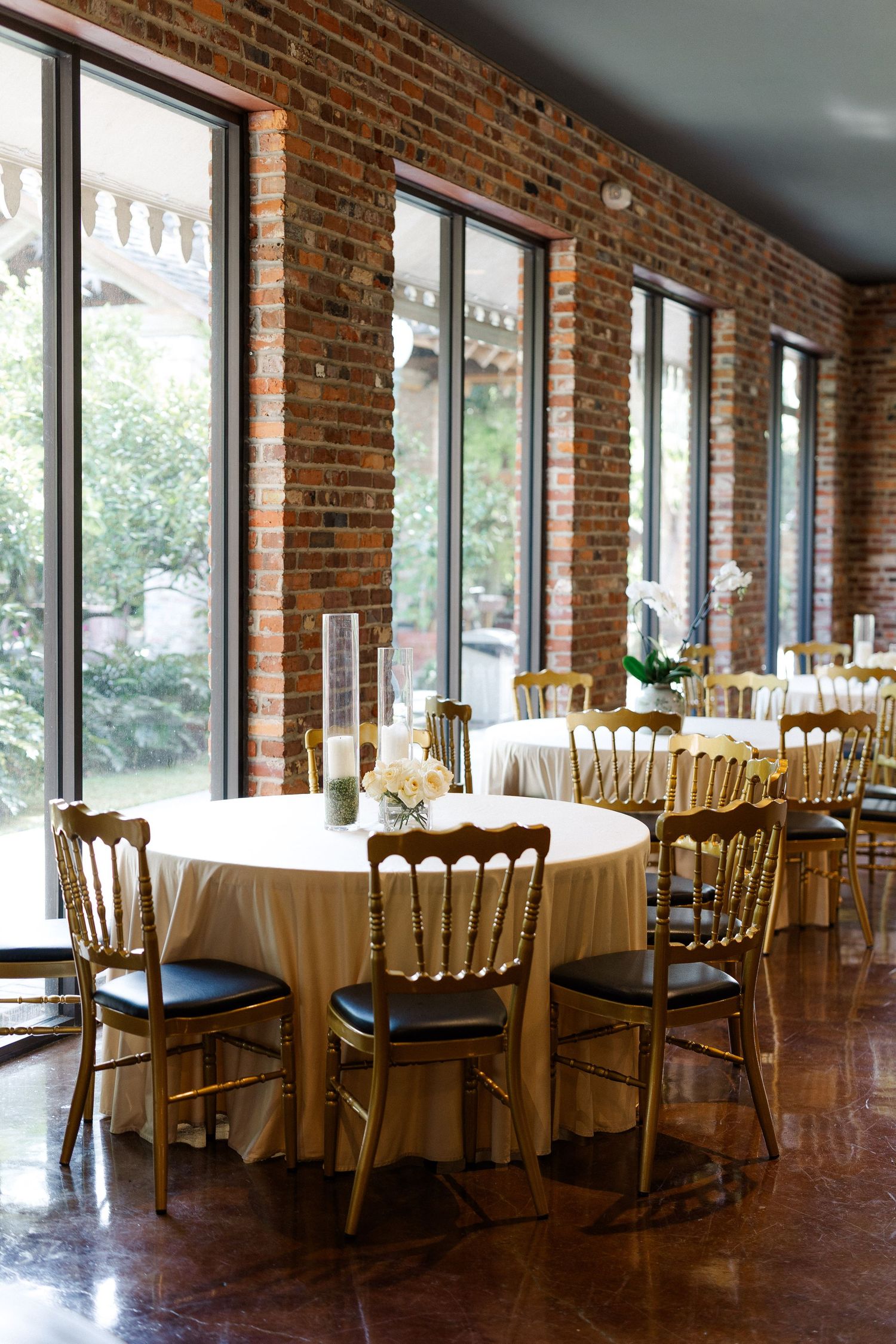 Elegant wedding reception venue with gold Chiavari chairs, round tables with ivory linens, and exposed brick walls with large windows.