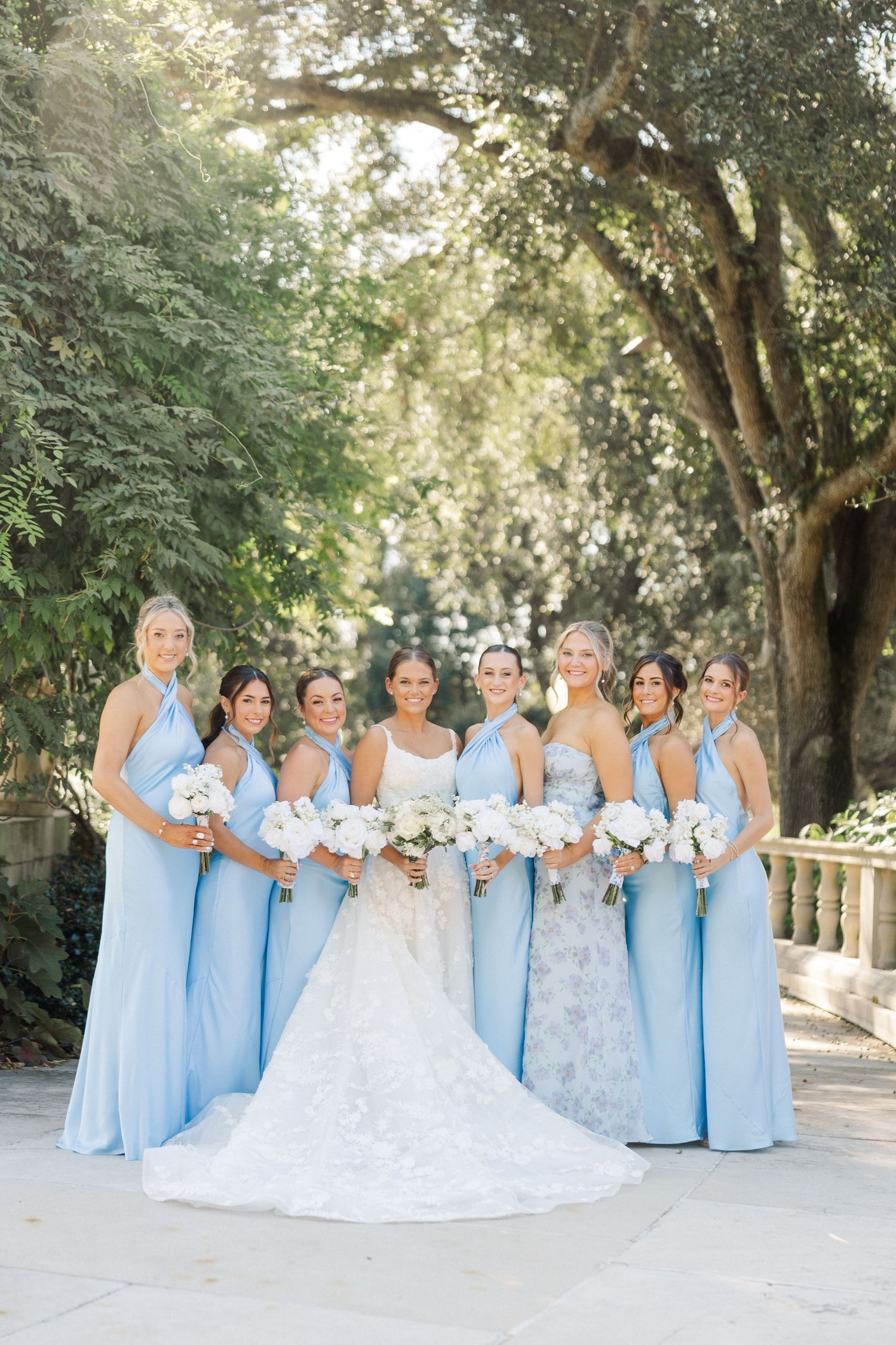A wedding party in light blue bridesmaid dresses and white bridal gown pose together outdoors under shady trees.