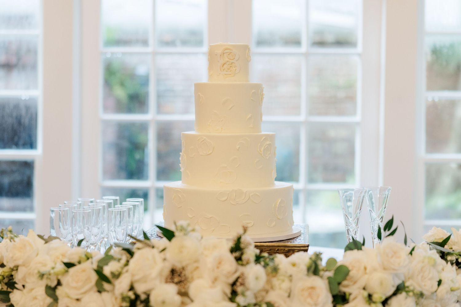 White wedding cake decorated with fresh flowers and greenery sits on an elegant display table near bright windows.