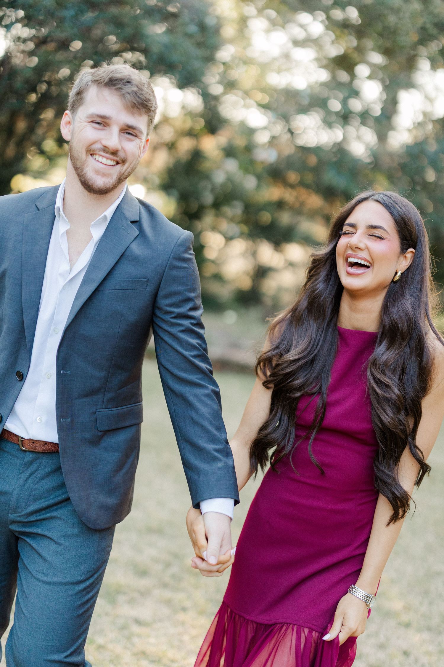 A couple laughs together outdoors in formal attire, with the woman in a burgundy dress and man in a navy suit.