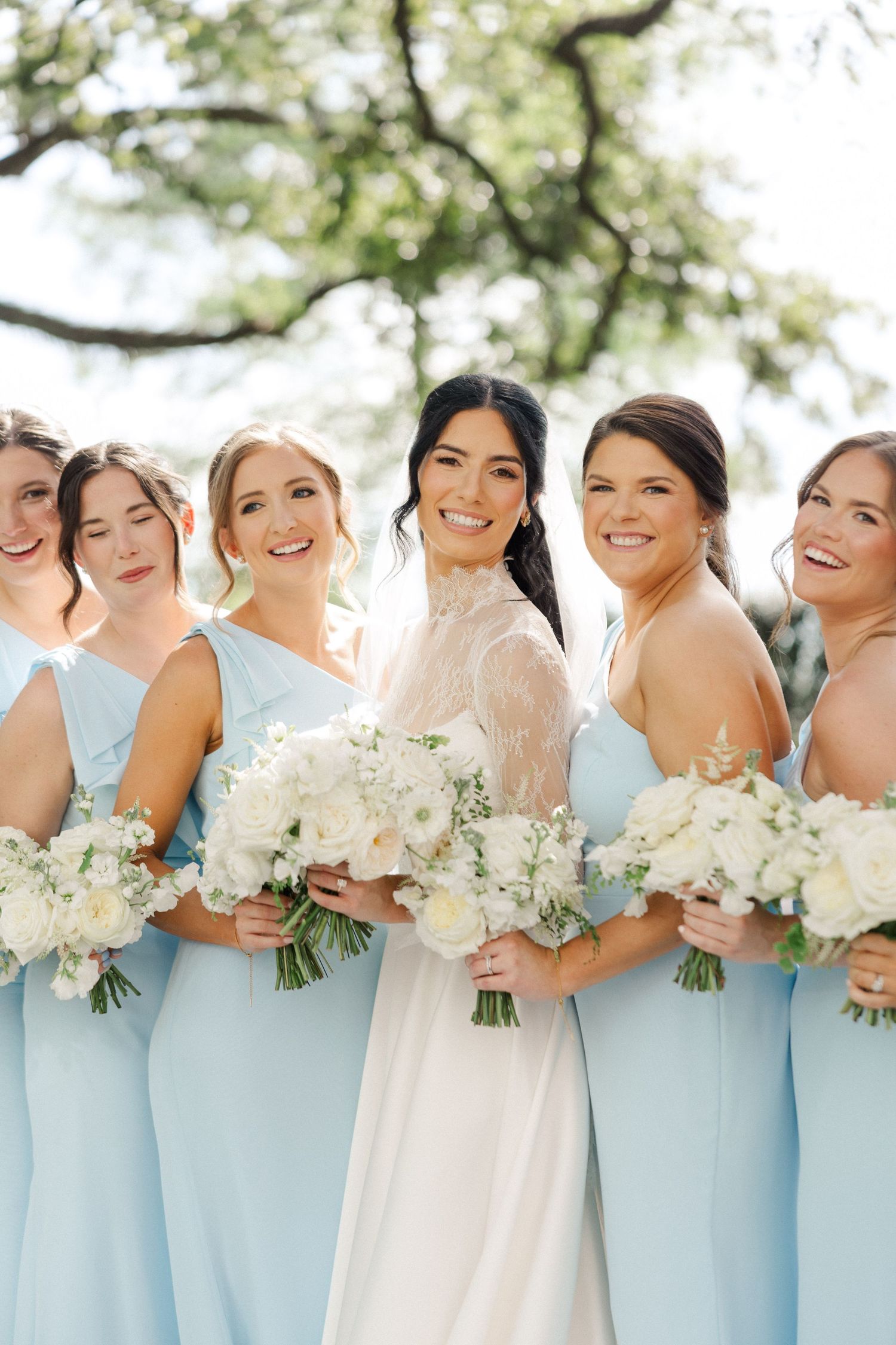 Bride in white gown with bridesmaids in light blue dresses holding white floral bouquets outdoors.